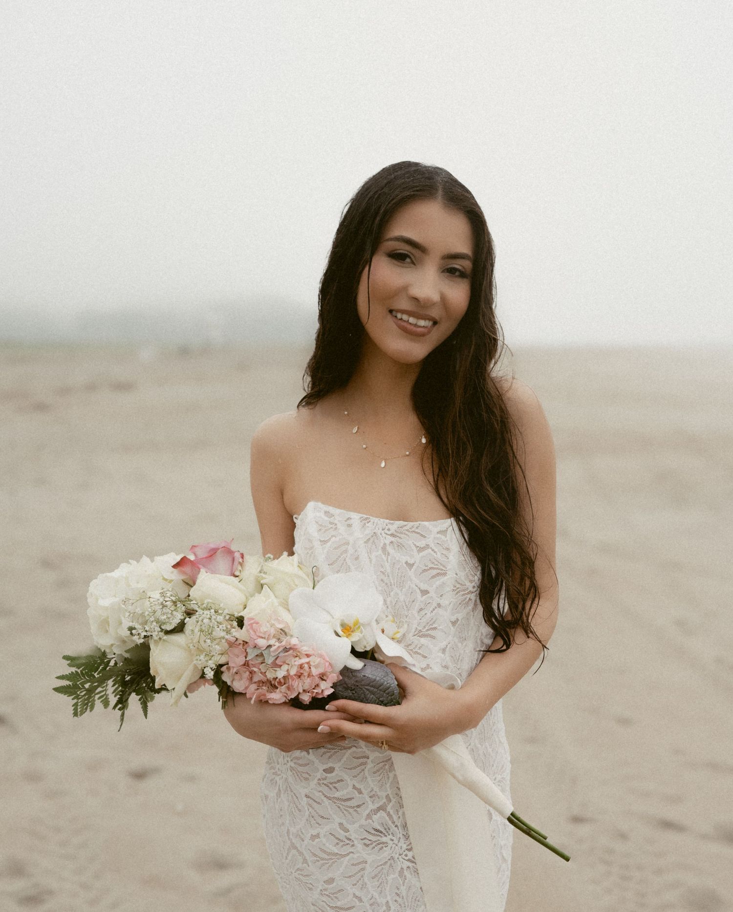 A bride in a lace wedding dress holds a bouquet while standing on a misty beach.