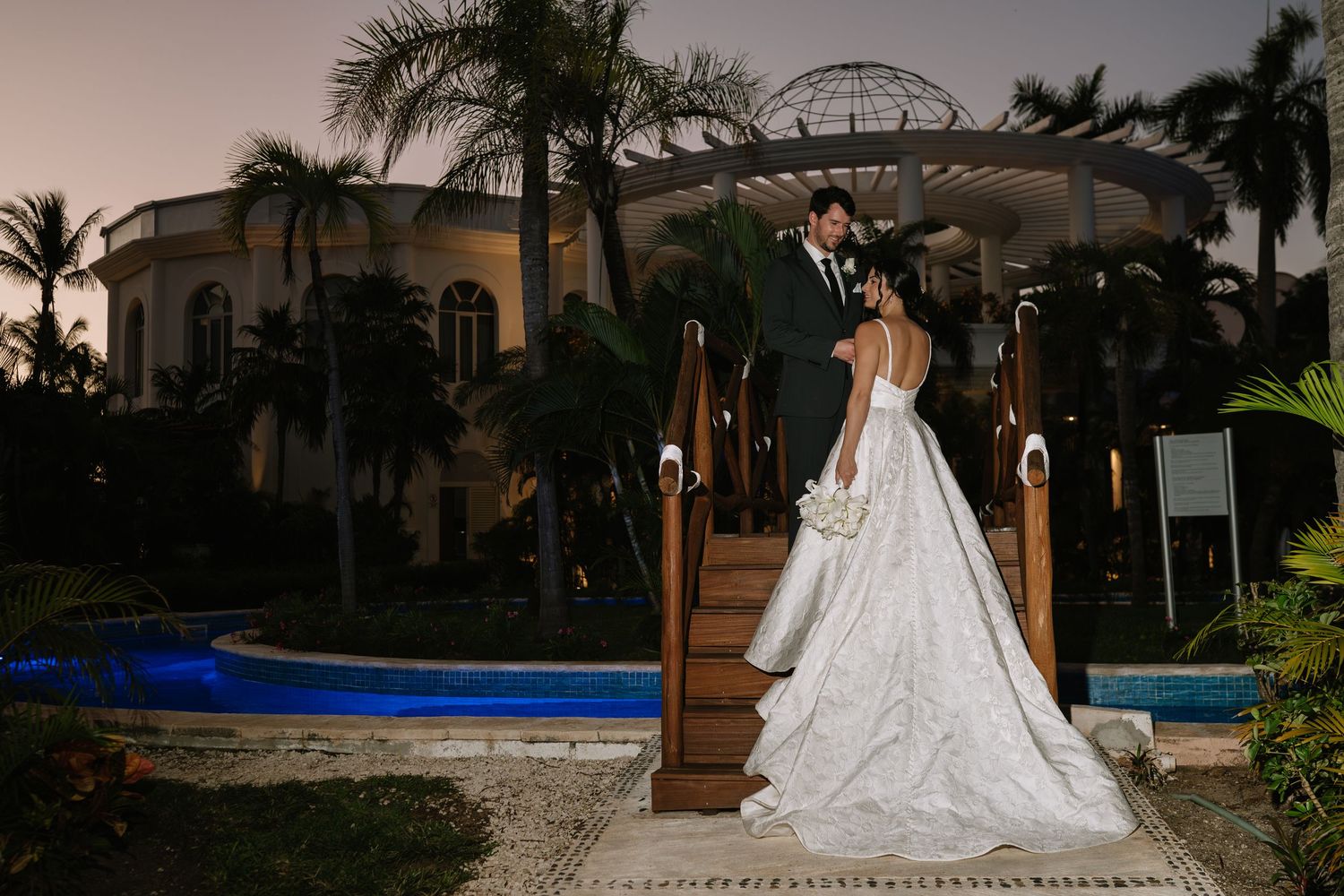 A long white wedding dress with a flowing train captured in a garden setting at dusk with palm trees in the background.