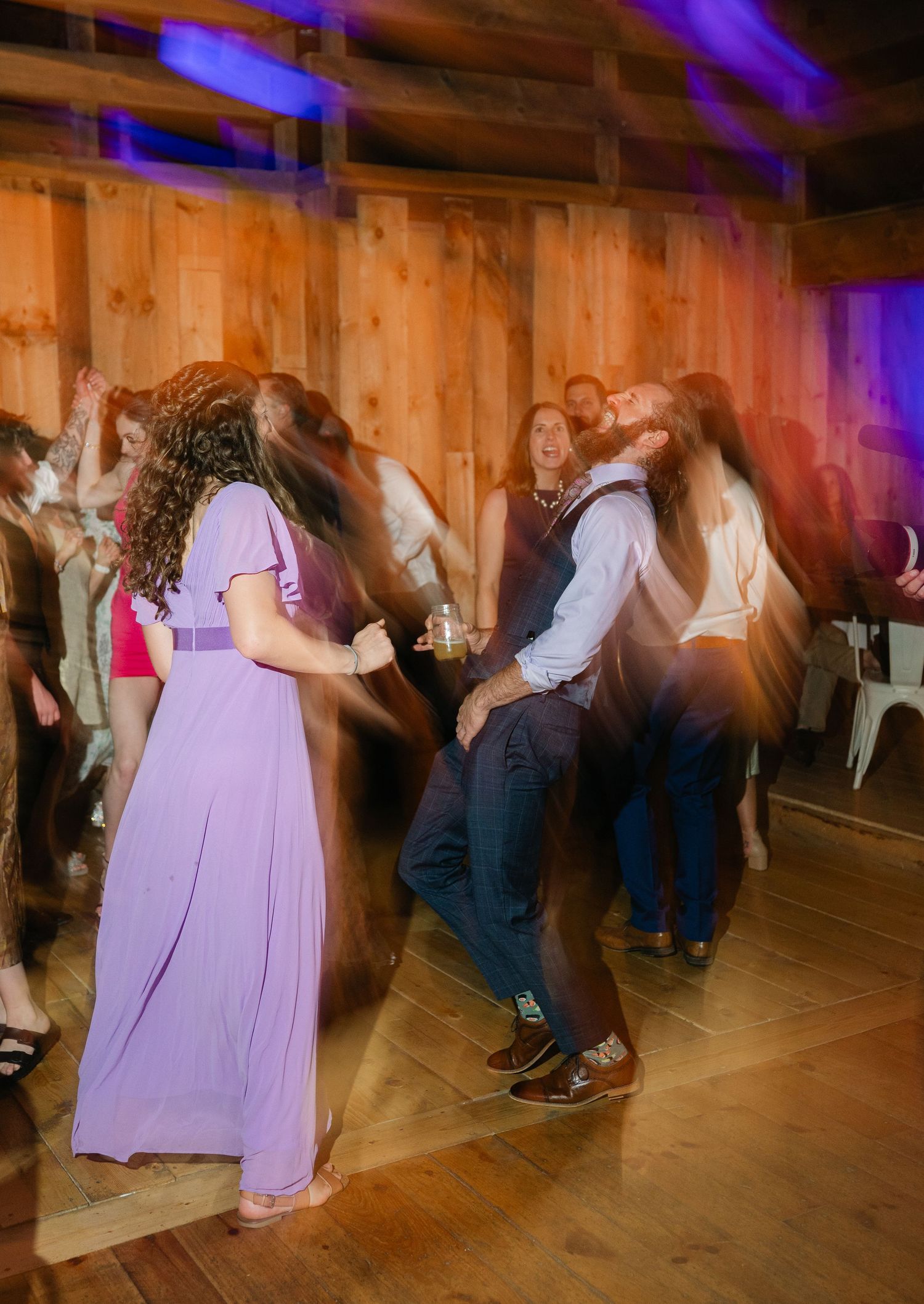 Guests in formal attire dancing at a wedding reception in a barn with purple ambient lighting.