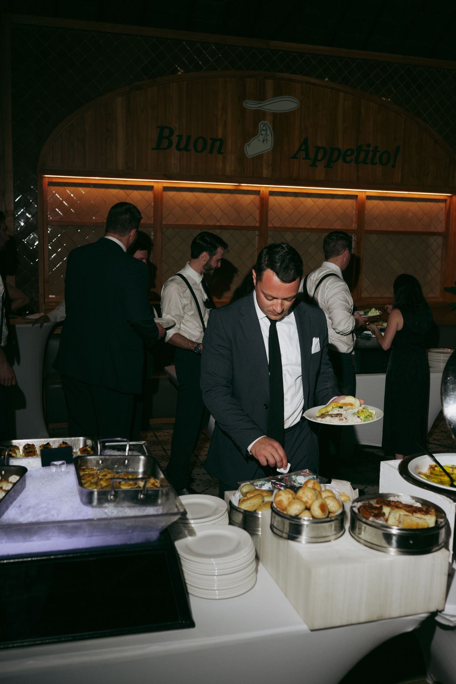 Wedding reception buffet line with servers in formal attire standing behind chafing dishes.