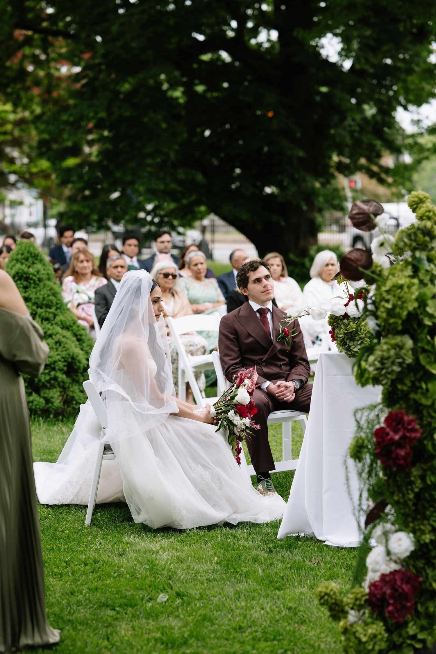 Bride in white gown and veil sits with groom in brown suit during outdoor wedding ceremony on green lawn.