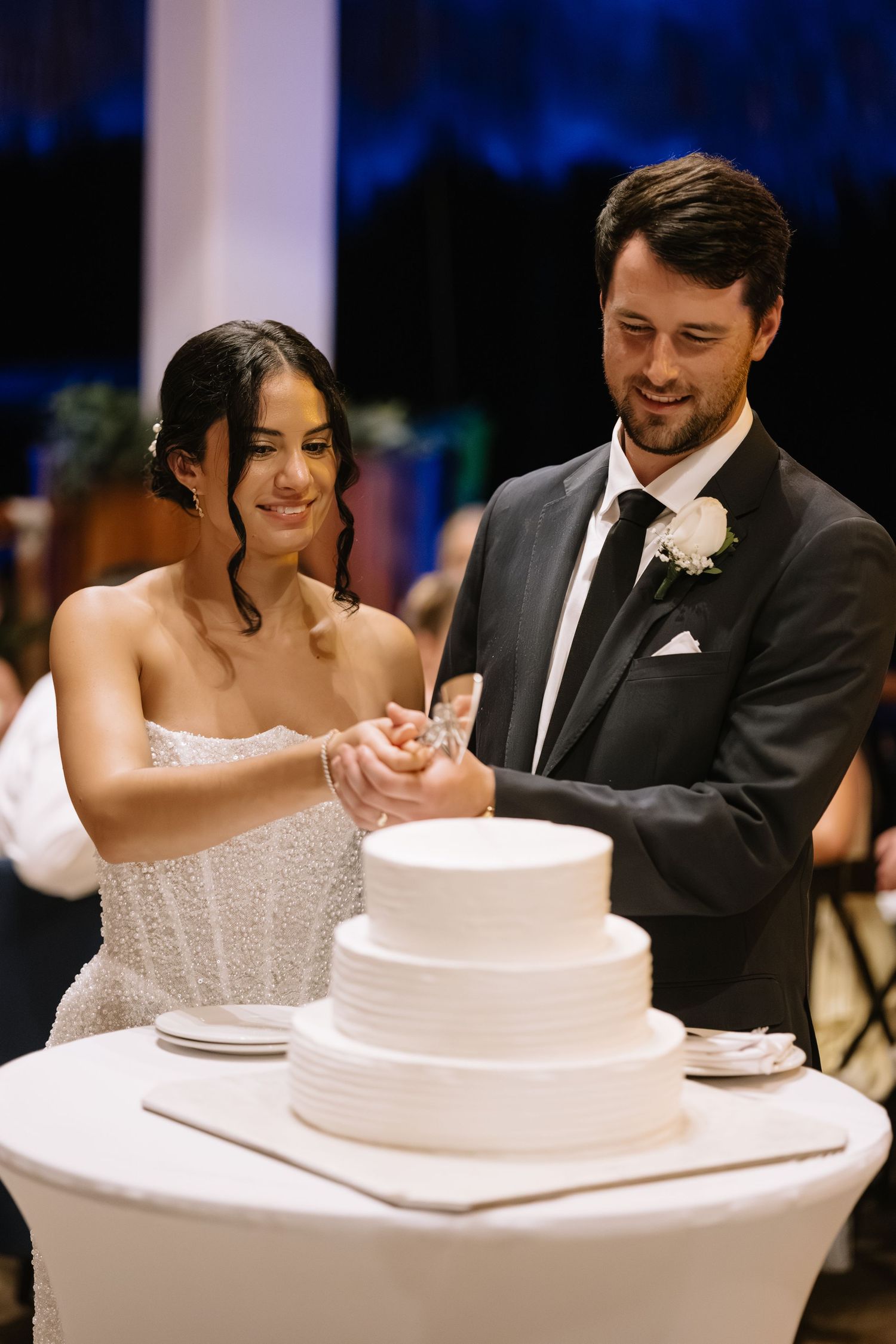 A wedding couple cuts into their elegant white tiered cake during their reception celebration.