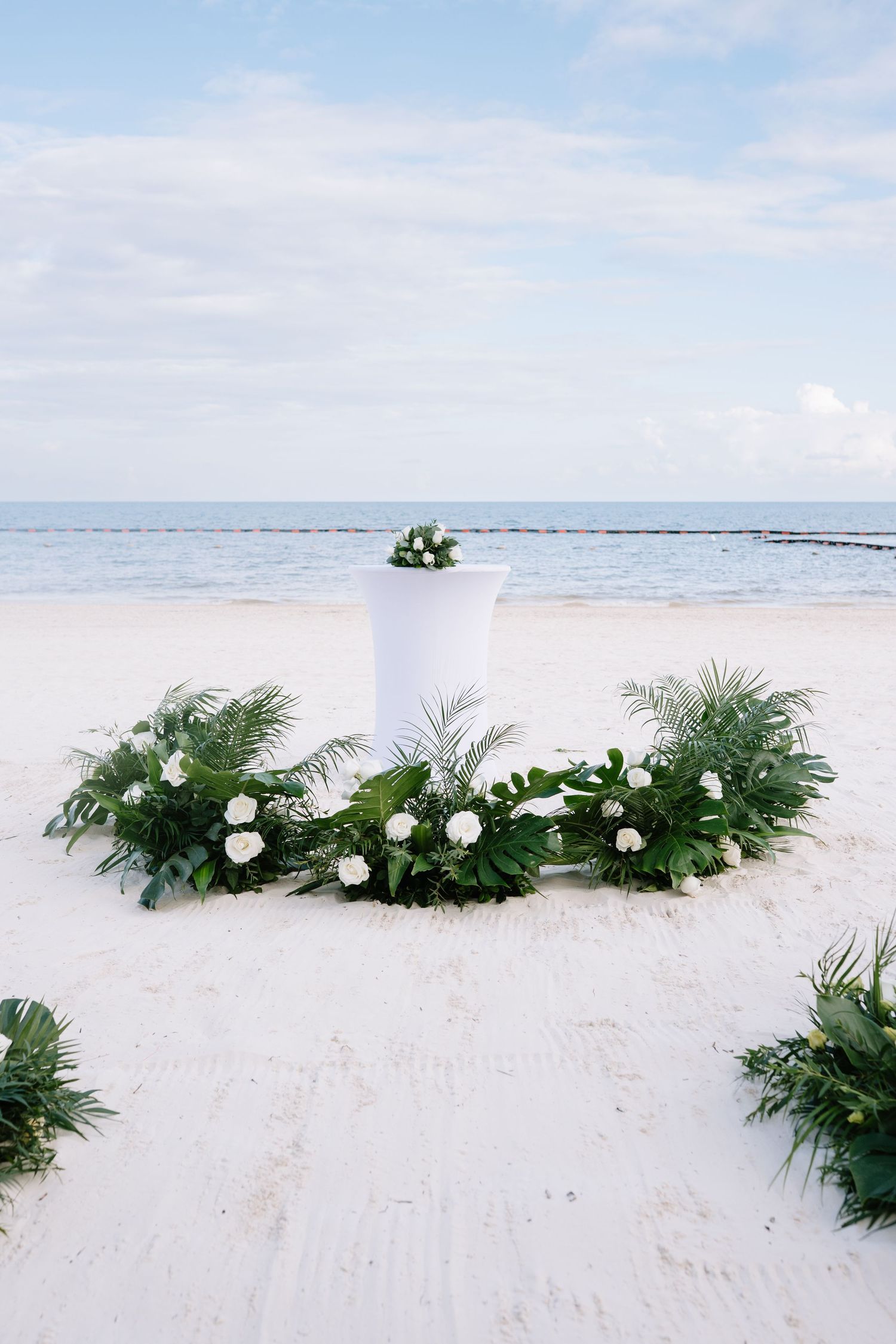 White floral arrangements and greenery decorate a pristine sandy beach with ocean views in the background.
