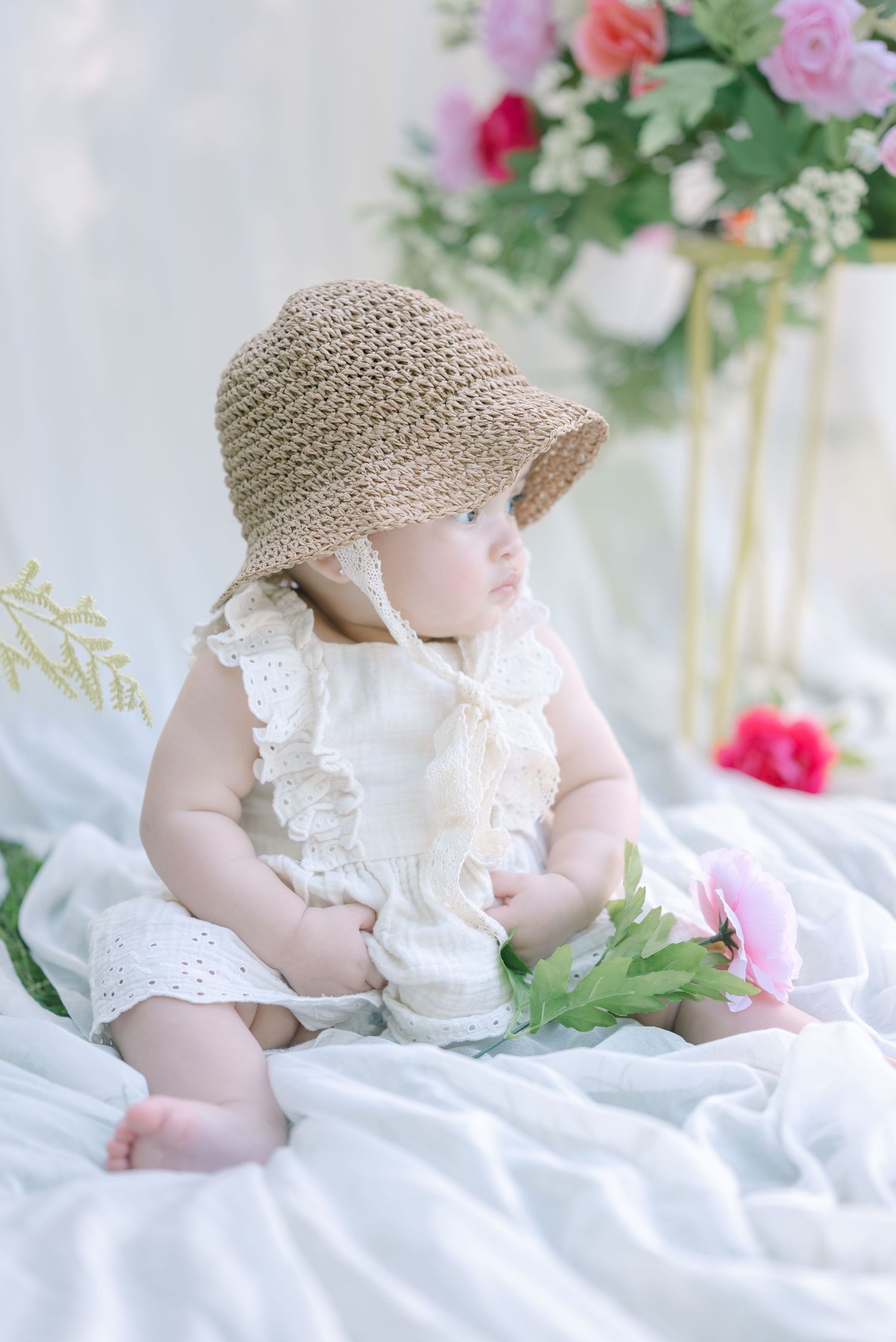 Baby in white lace dress and sun hat sitting among pink and purple flowers.
