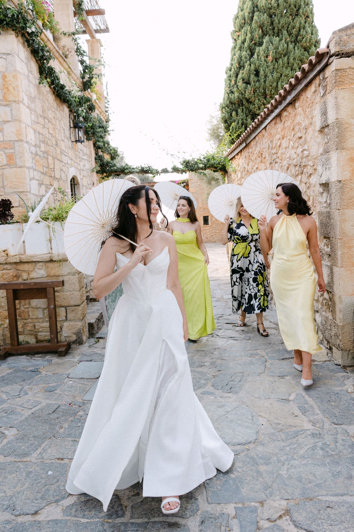 A bride and her friends holding sun umbrellas and walking candidly looking at each other in a farm in Crete