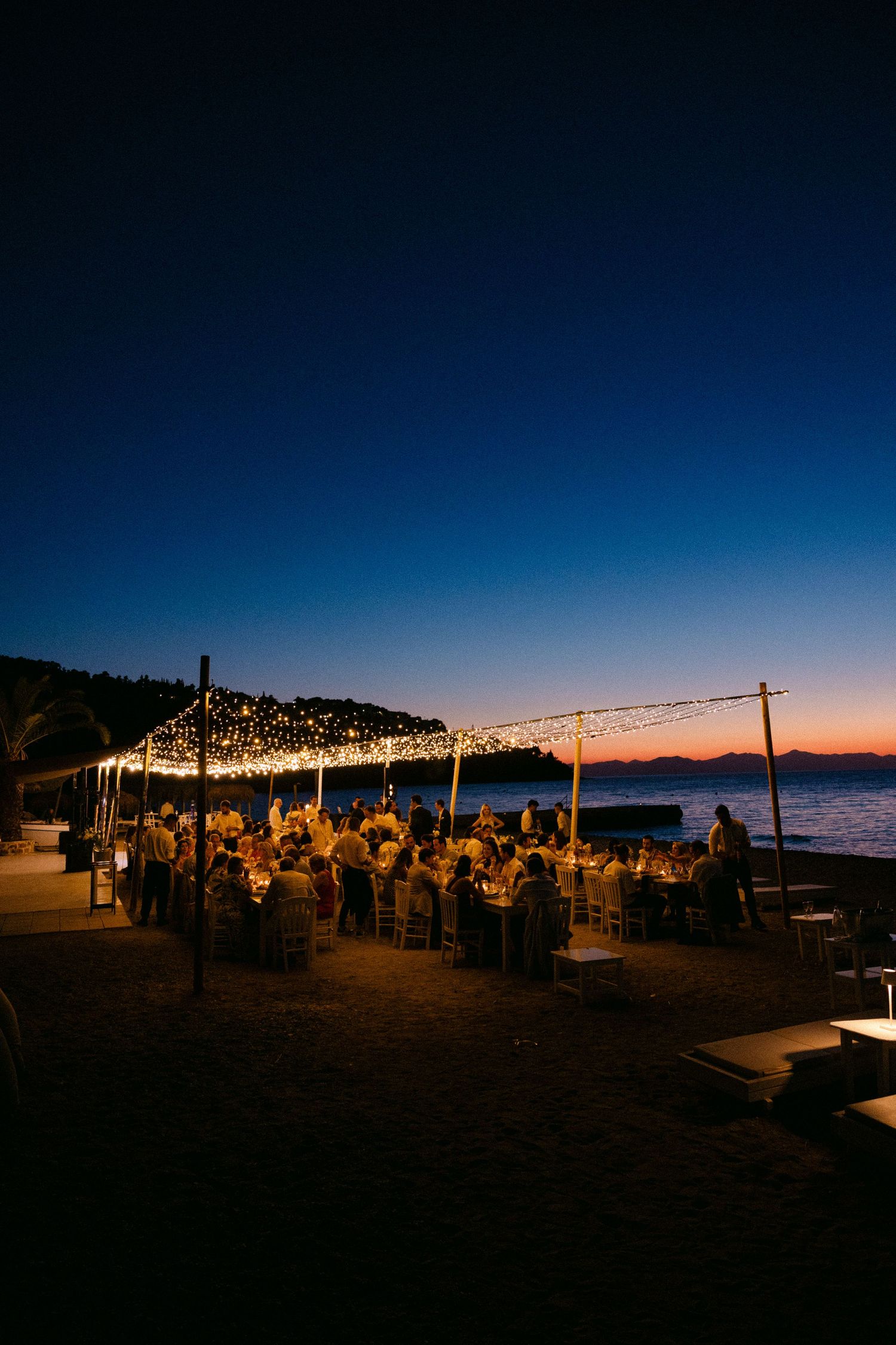 a wide photo of an open-air wedding dinner in a beach bar in spetses - there are three wide tables under carnival lights at blue hour