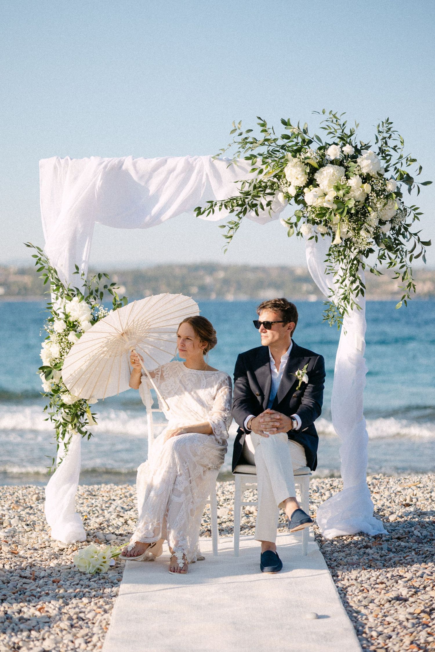 a bride and groom sitting in chairs under a drape arch with white flowers during their beach wedding ceremony
