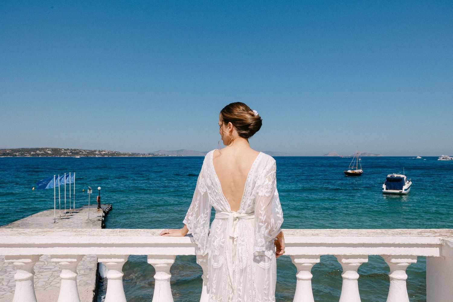 a bride with an up hair do and a lace long-sleeve wedding dress standing in a balcony with the sea as a background in a sunny setting