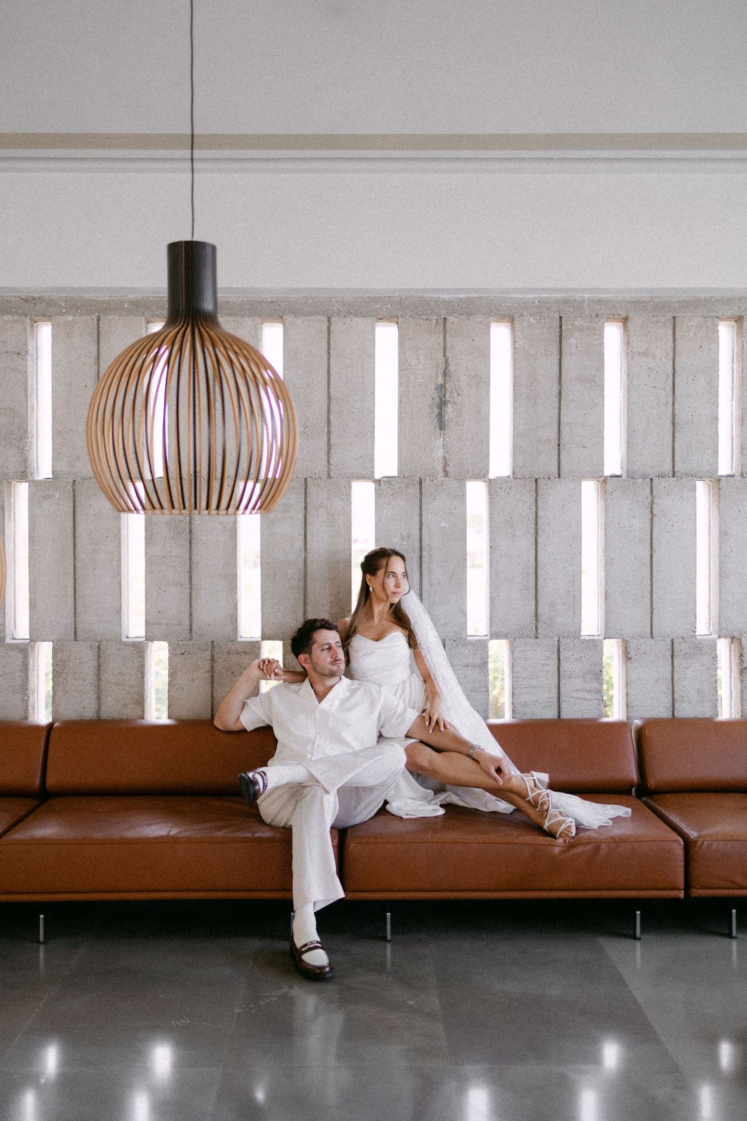 a bride and groom sitting in a brown leathered sofa in an industrial setting holding hands and looking to the right in an editorial pose