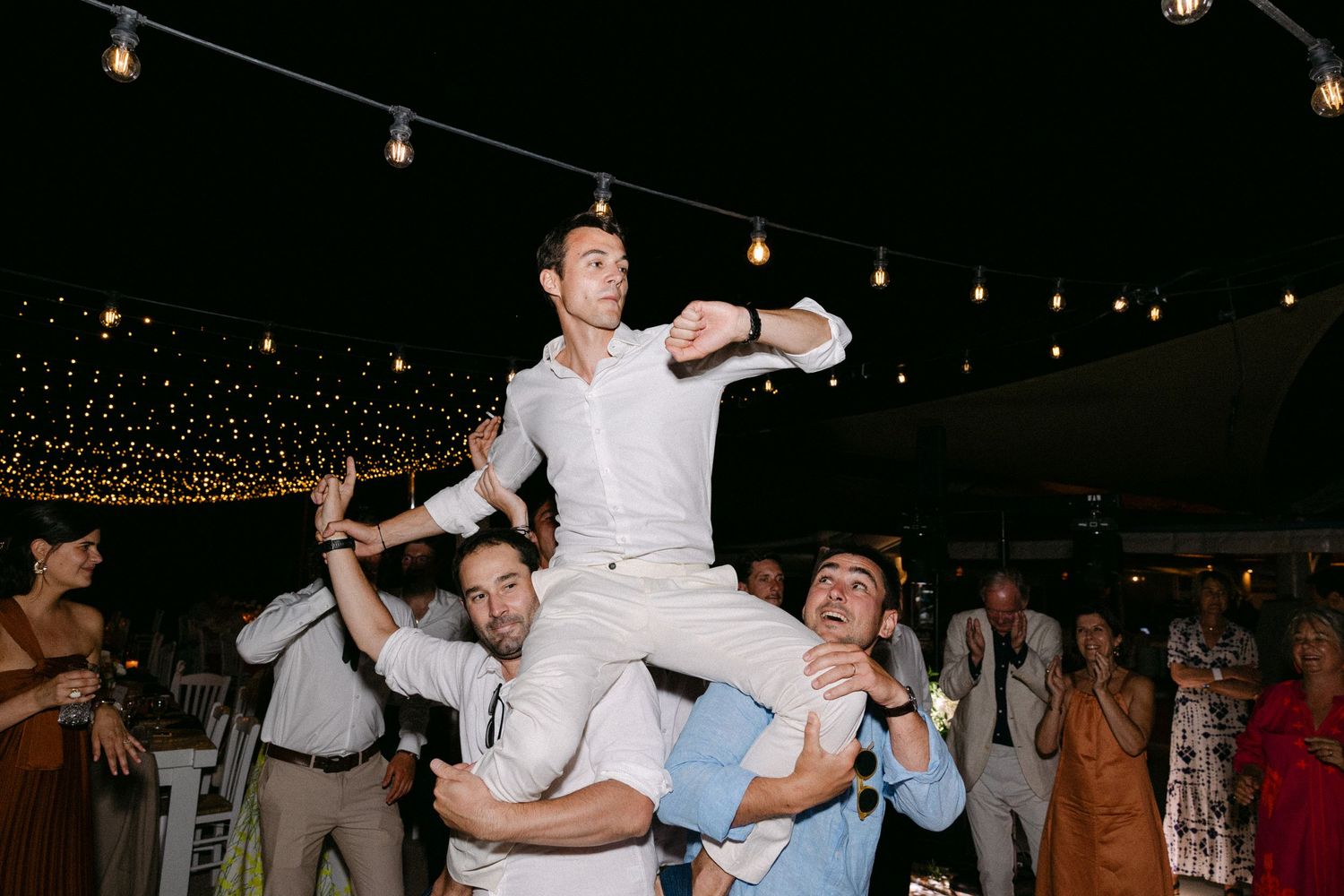 the groom sitting in the shoulders of two guests during a wedding party, dancing happily