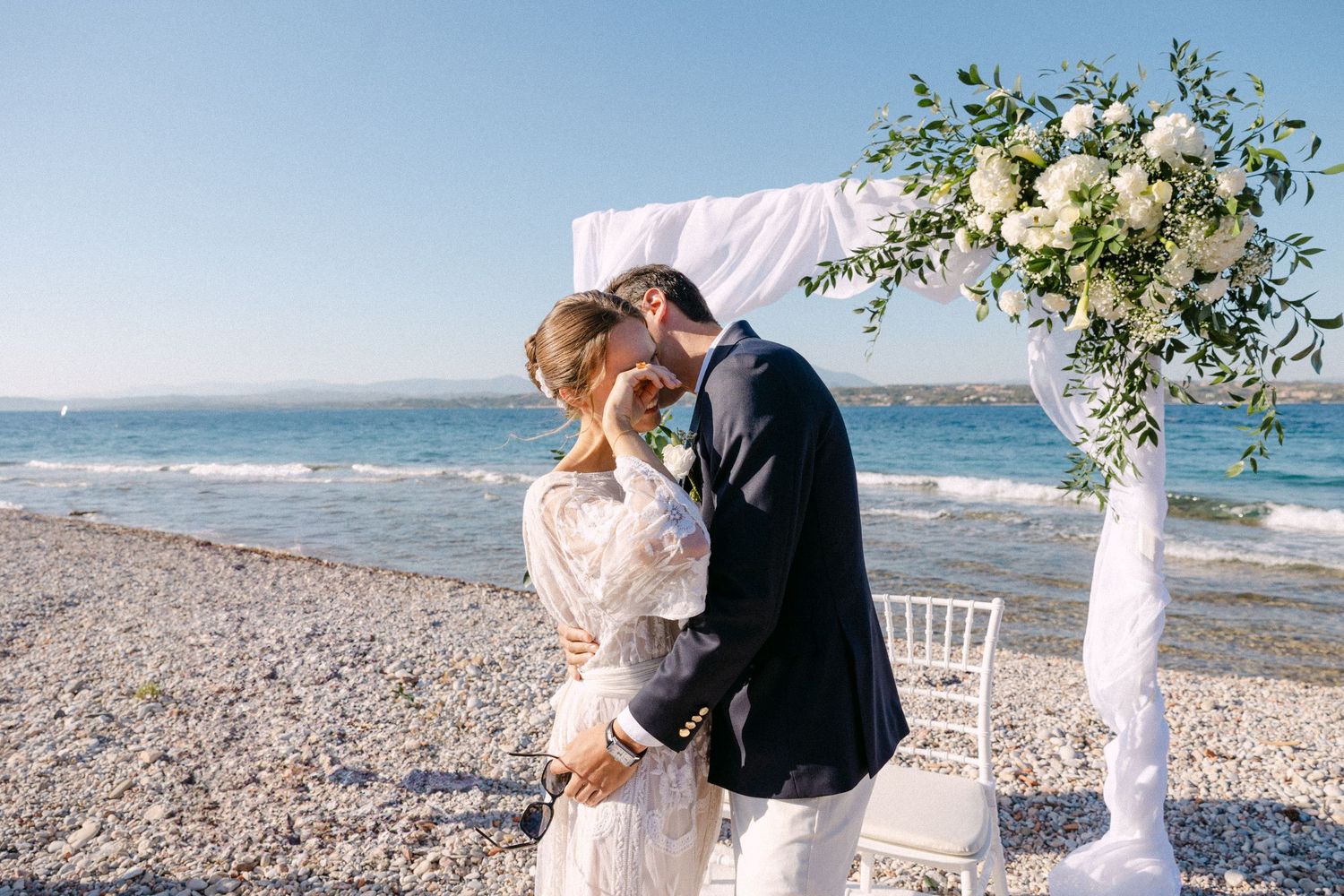 a groom hugging his bride while she is emotionally crying - the background is a white drape arch with white flowers on the beach