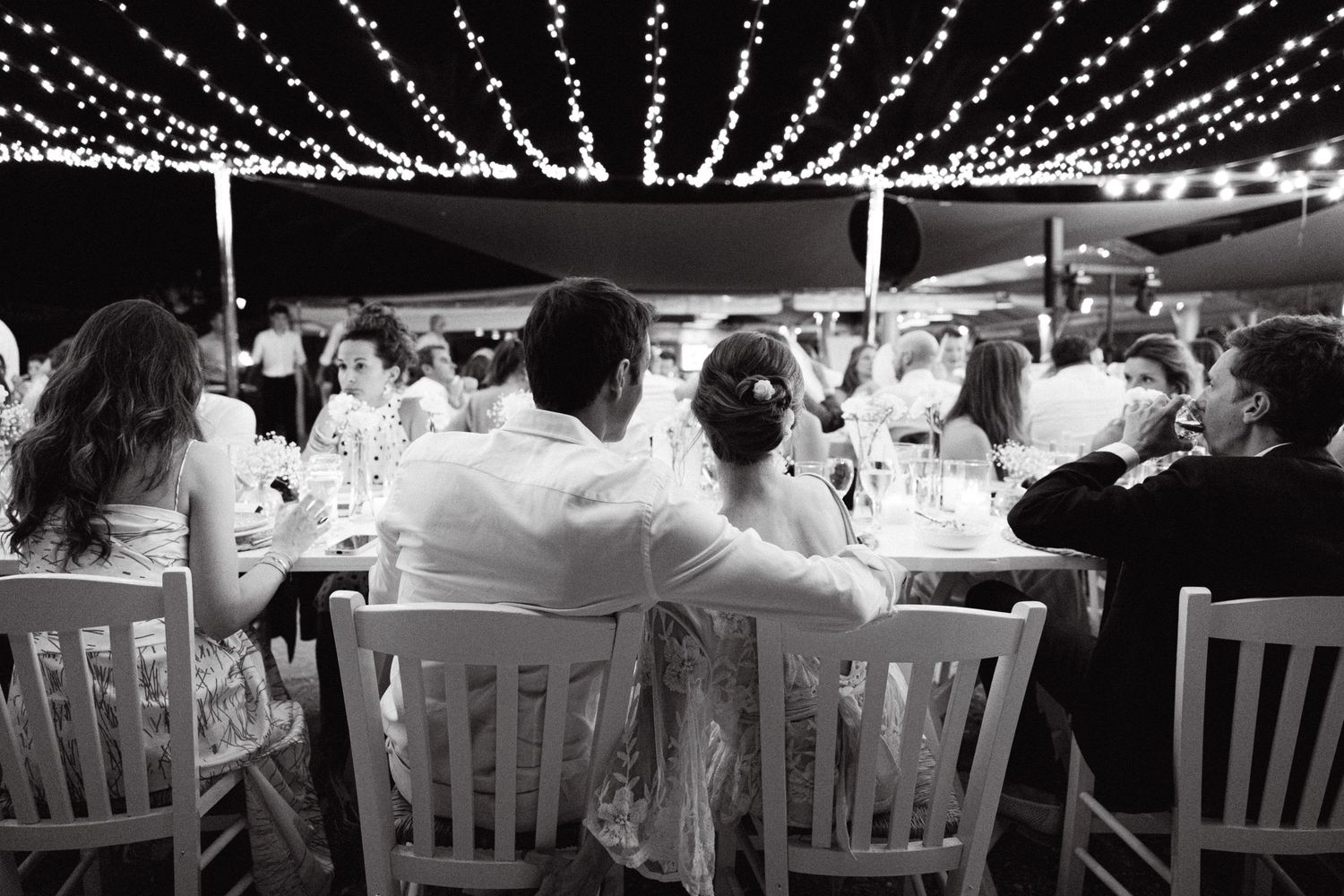 a black and white photo of newlyweds with their backs on the camera hugging at the dinner table of their wedding
