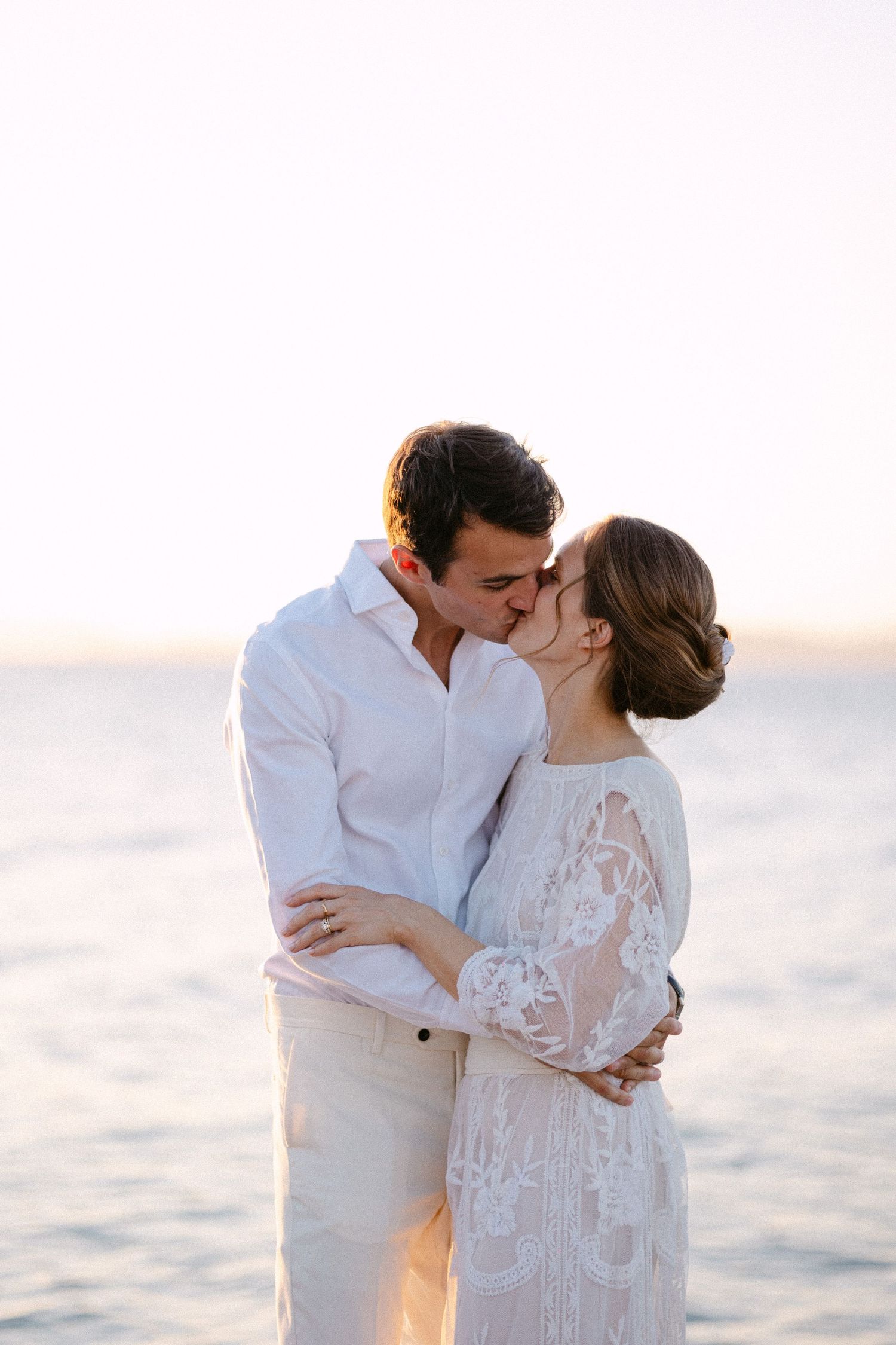 a bride and groom hugging and kissing each other at sunset with the sea in the background
