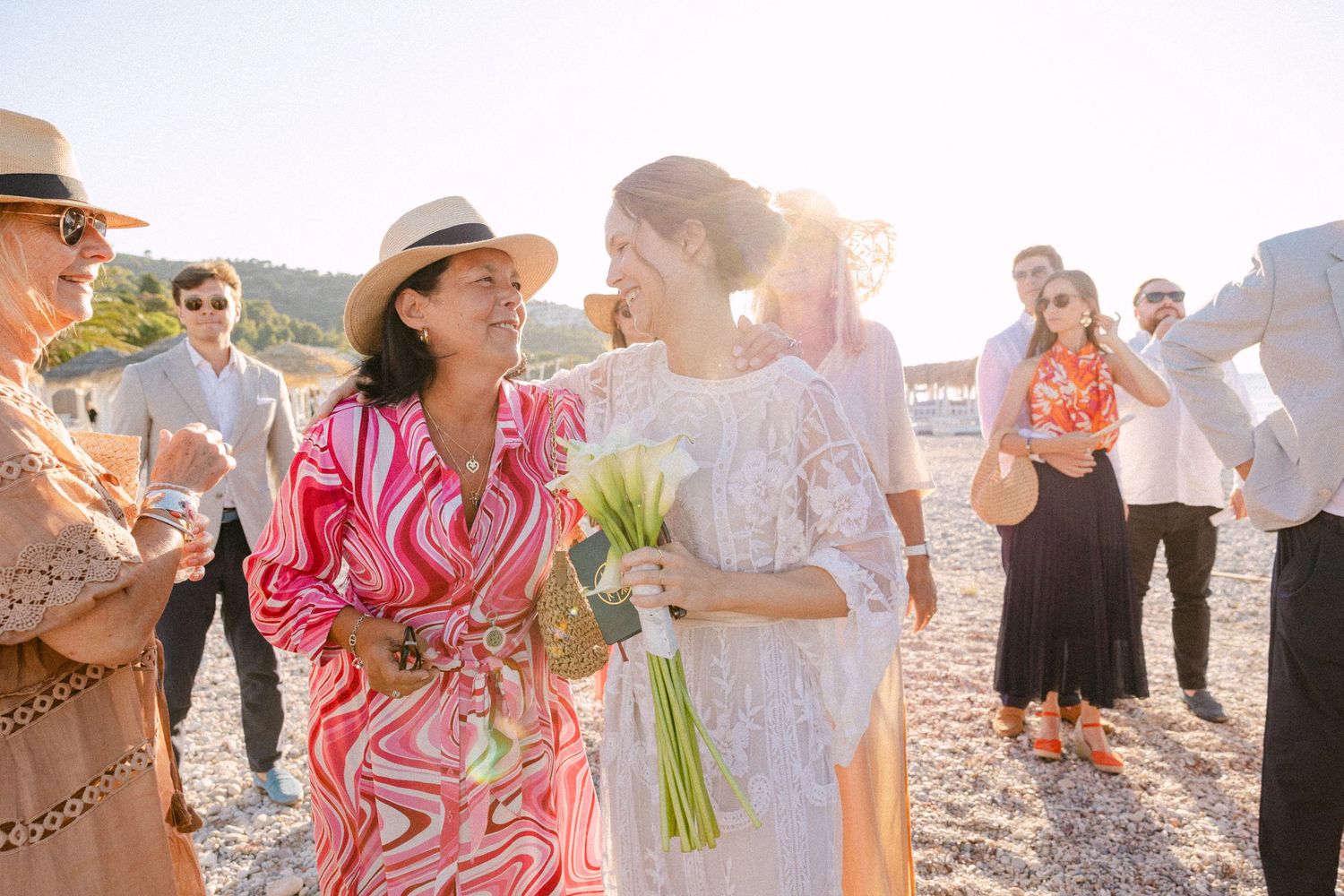 a bride hugging a guest among other guests at a beach at afternoon, holding a white Lillies bouquet