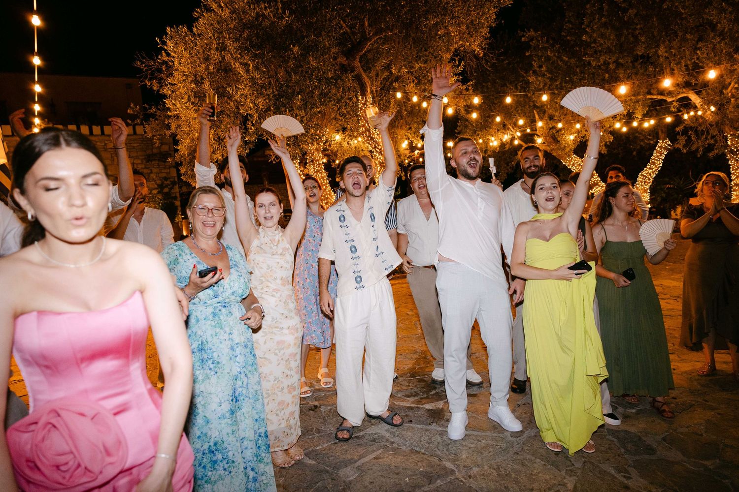 wedding guests cheering with hands up in the air in an outdoors wedding reception at night, some of them holding fans