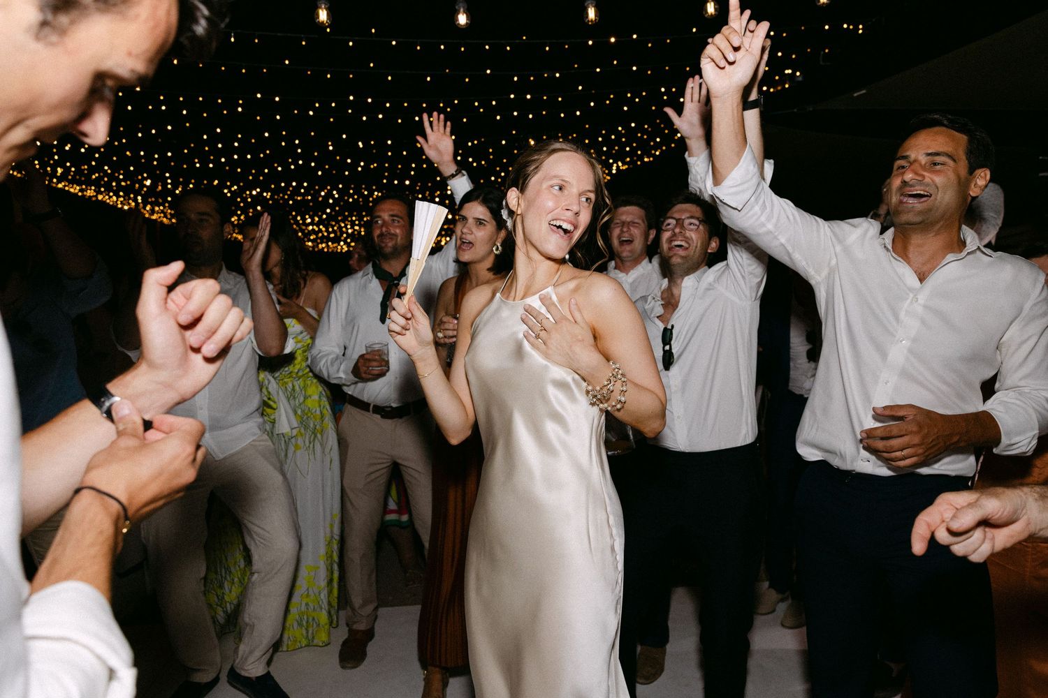 a bride holding a fan and standing among guests that sign loudly
