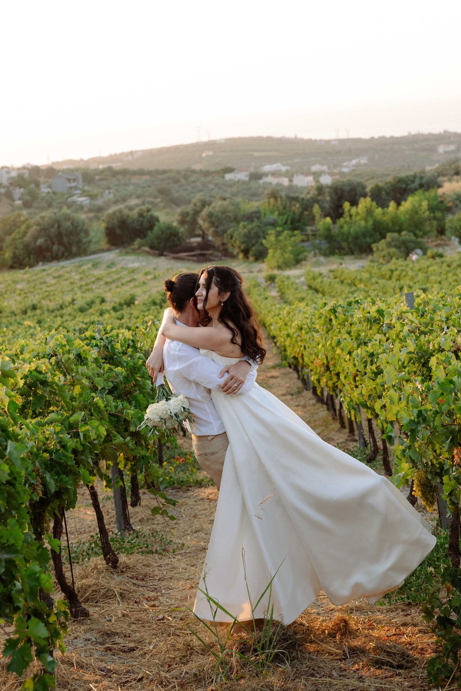 a groom holding and twirling a bride while she laughs holding her white flowers bouquet in a vineyard at sunset