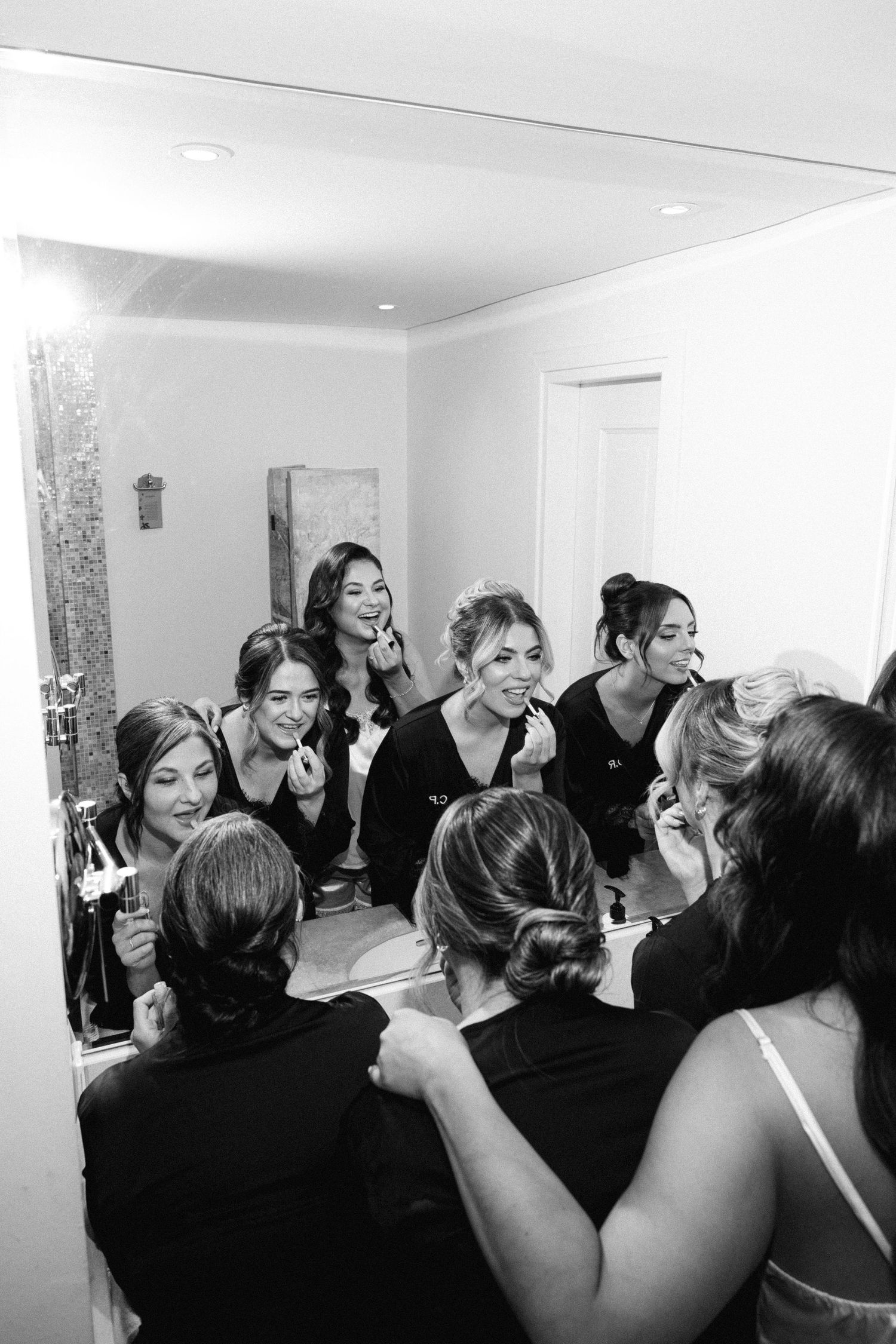 A black and white photo showing a bride and her bridal party in their pyjamas putting on lipstick in front of a mirror during getting ready