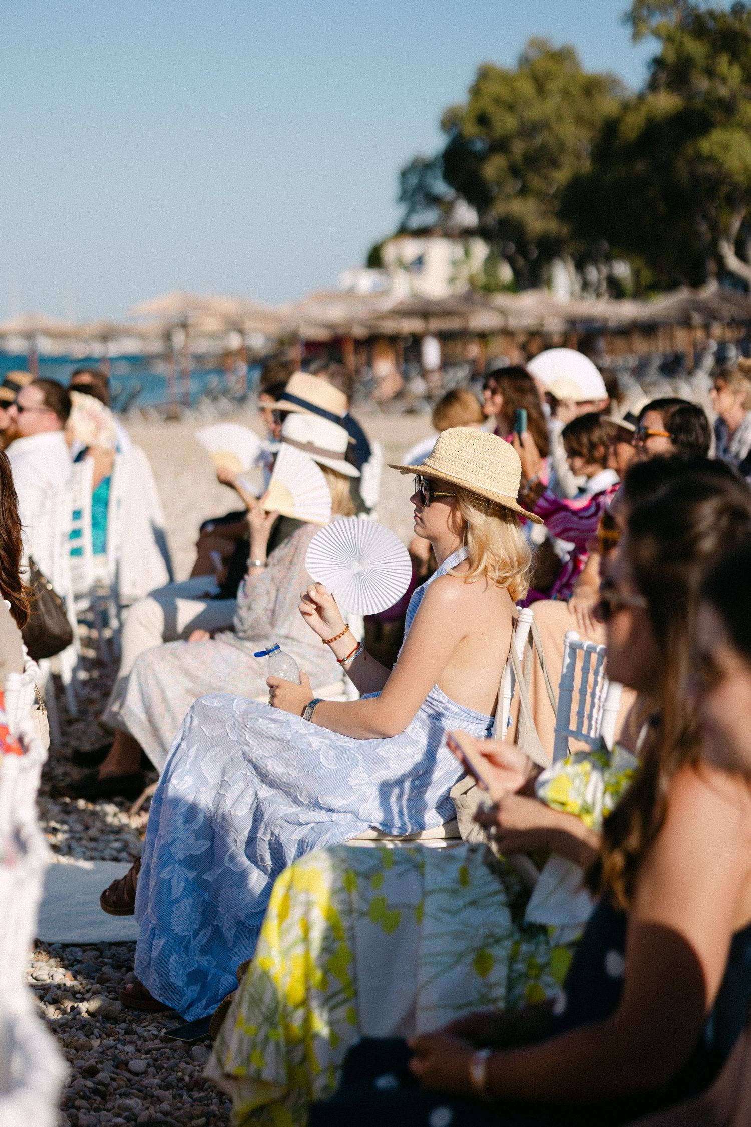 wedding guests sitting in chairs during a sunny open-air wedding ceremony, using fans and wearing hats