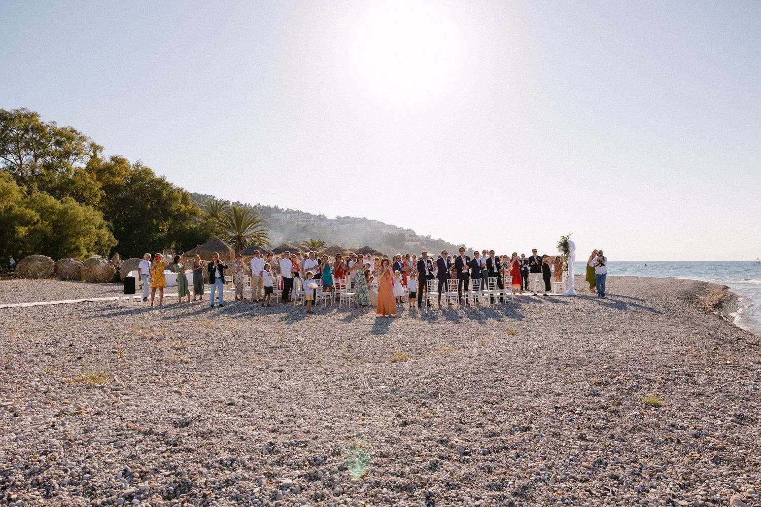 a wide photo of wedding guests during a sunny open-air beach wedding ceremony clapping