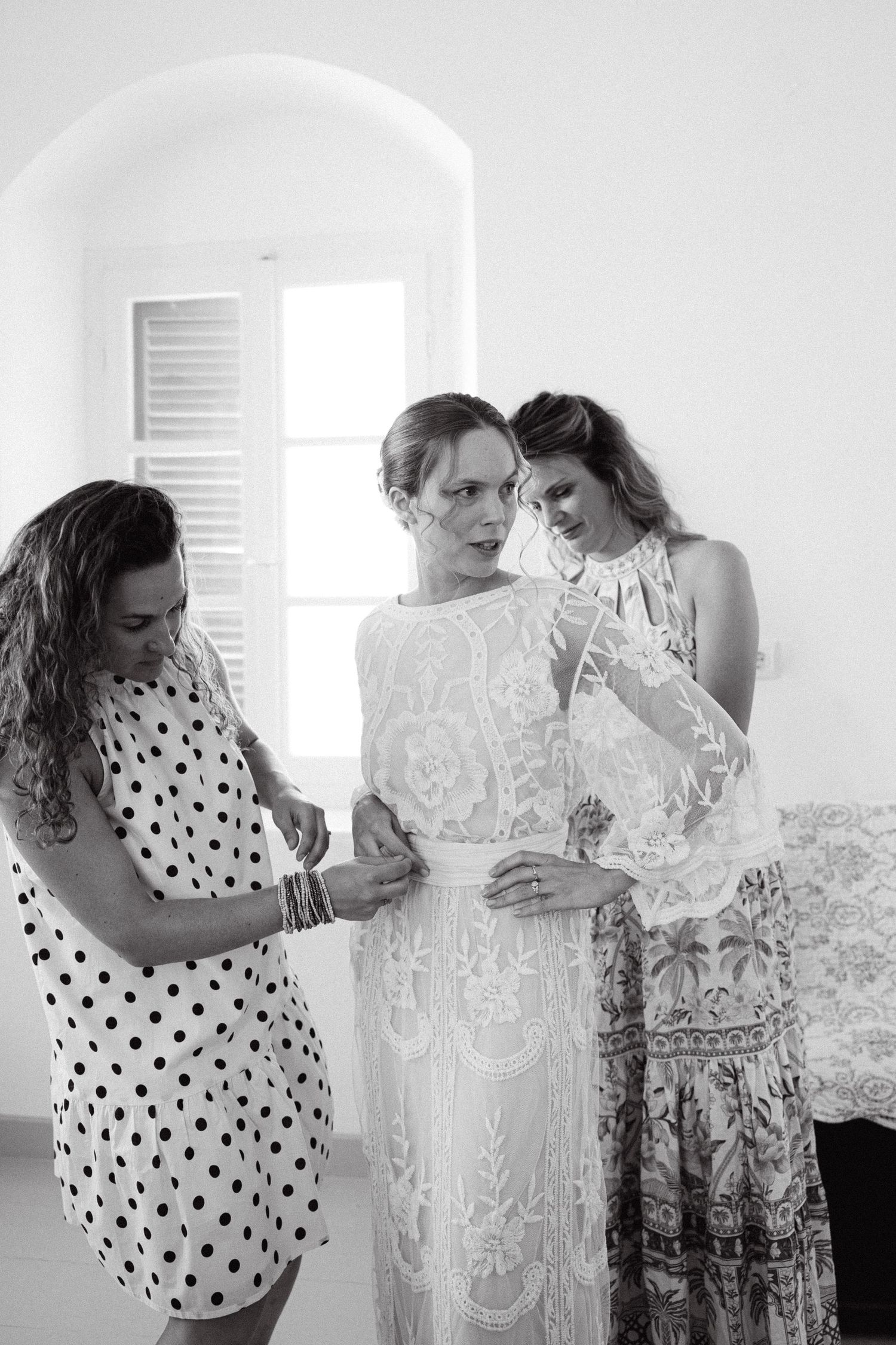 a black and white photo of a bride in a white room being dressed by her two friends into her long-sleeved lace wedding dress