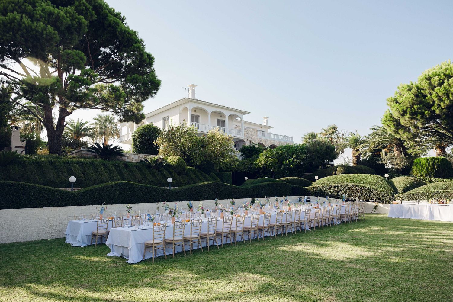 A wedding reception outdoors garden area with a villa in the background showing 2 long tables decorated with flowers in Athens Greece