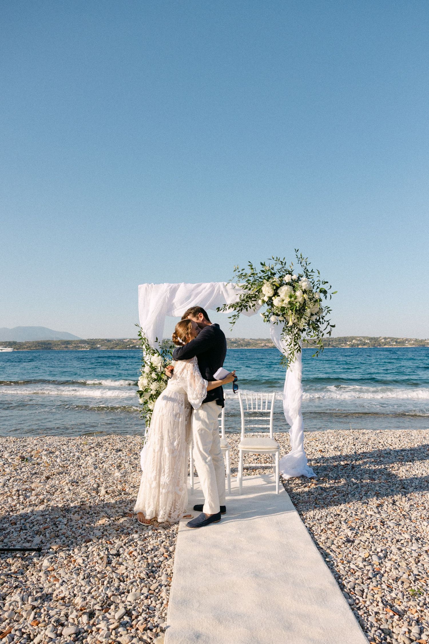 a bride and groom hugging and kissing under a drape arch with white flowers during their beach wedding ceremony in spetses