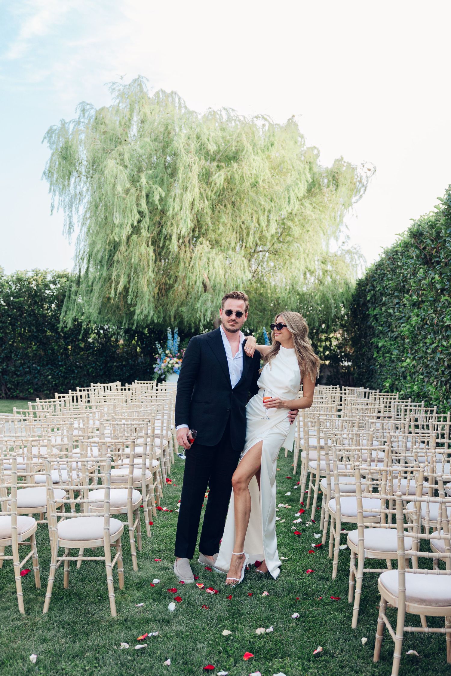 A bride and groom posing like fashion models wearing sunglasses in front of their wedding ceremony area surrounded by chairs and greenery