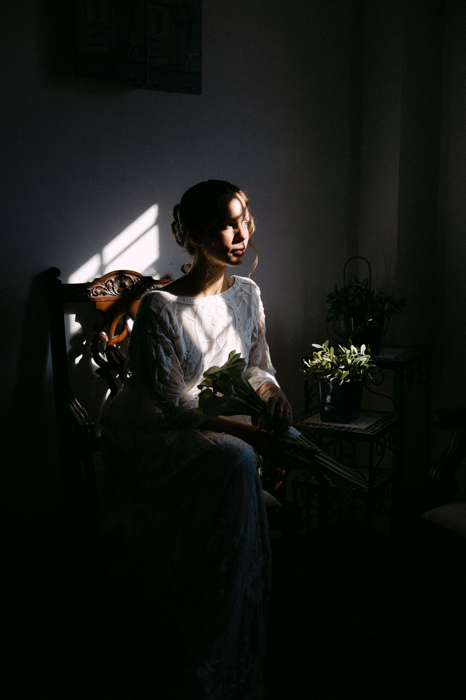 a moody portrait of a bride being dimly-lit from window light while sitting in an armchair, and looking outside the window