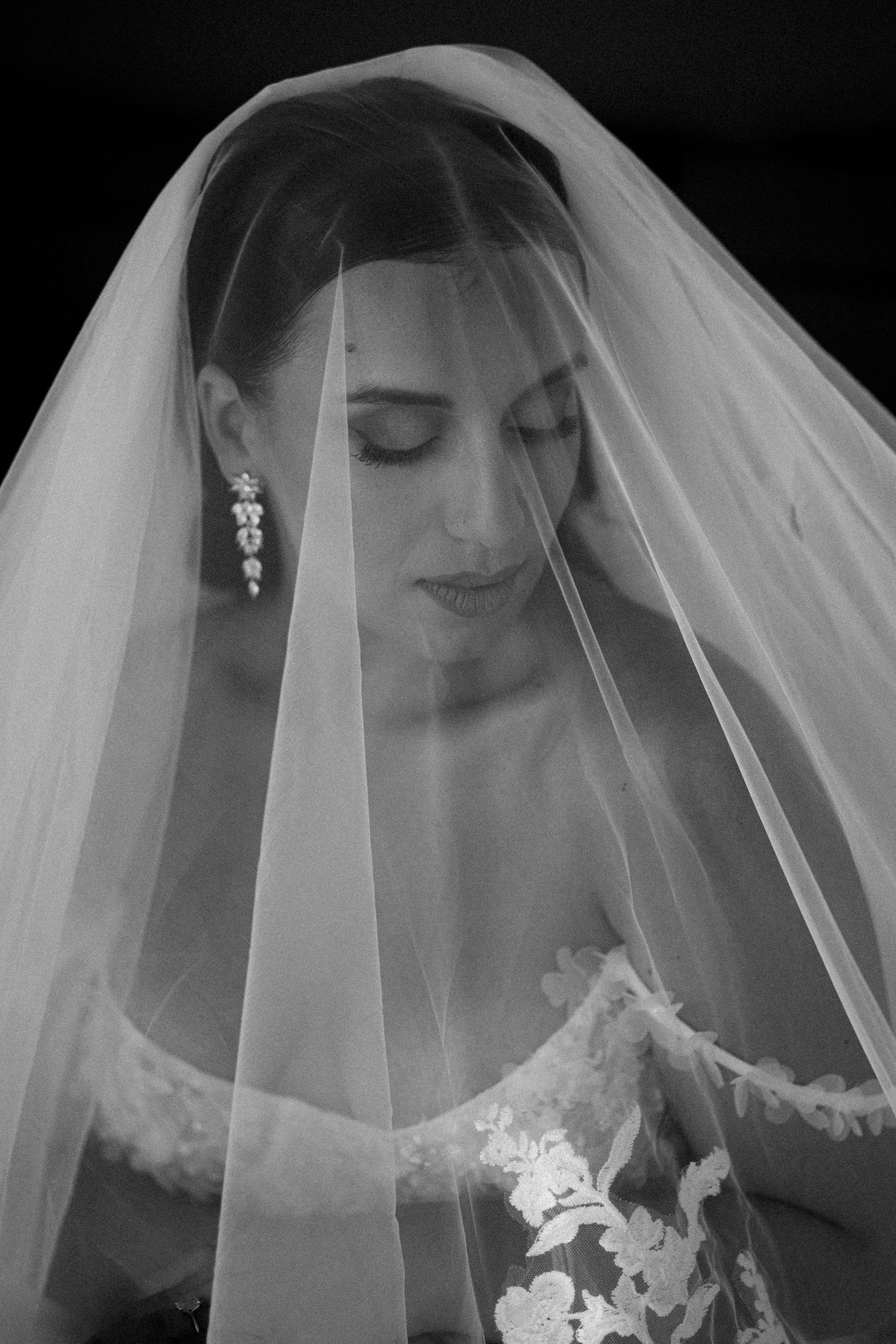 a black and white close up portrait of a bride under her veil smiling very softly and looking down, she is wearing crystal earrings