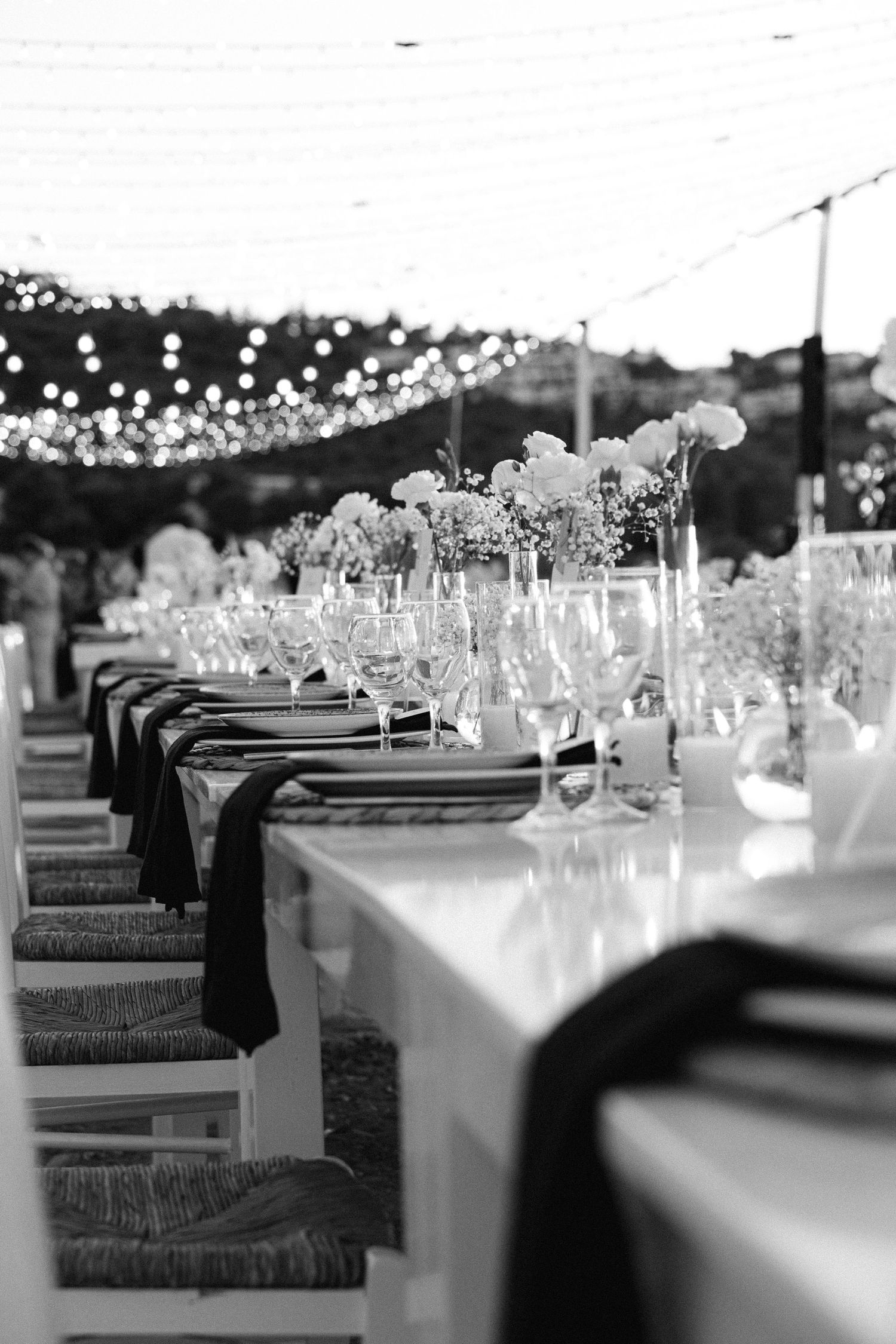 a black and white photo of wedding table decorations showing white flowers in a vase, lit candles, plates and glass wines