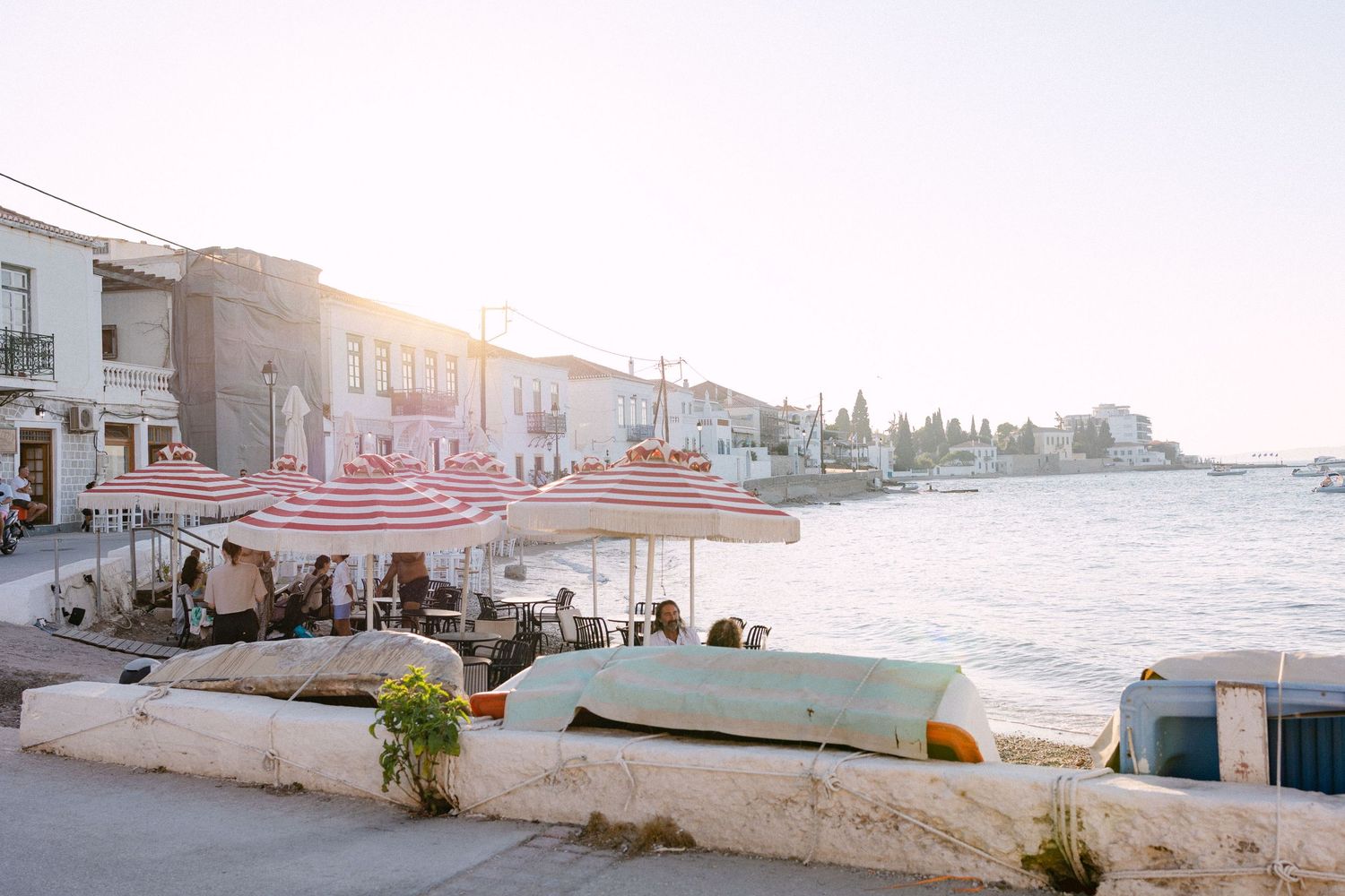 A beach in Spetses during golden hour while the sun is setting - we see red and white stripped umbrellas, boats, the sea and houses