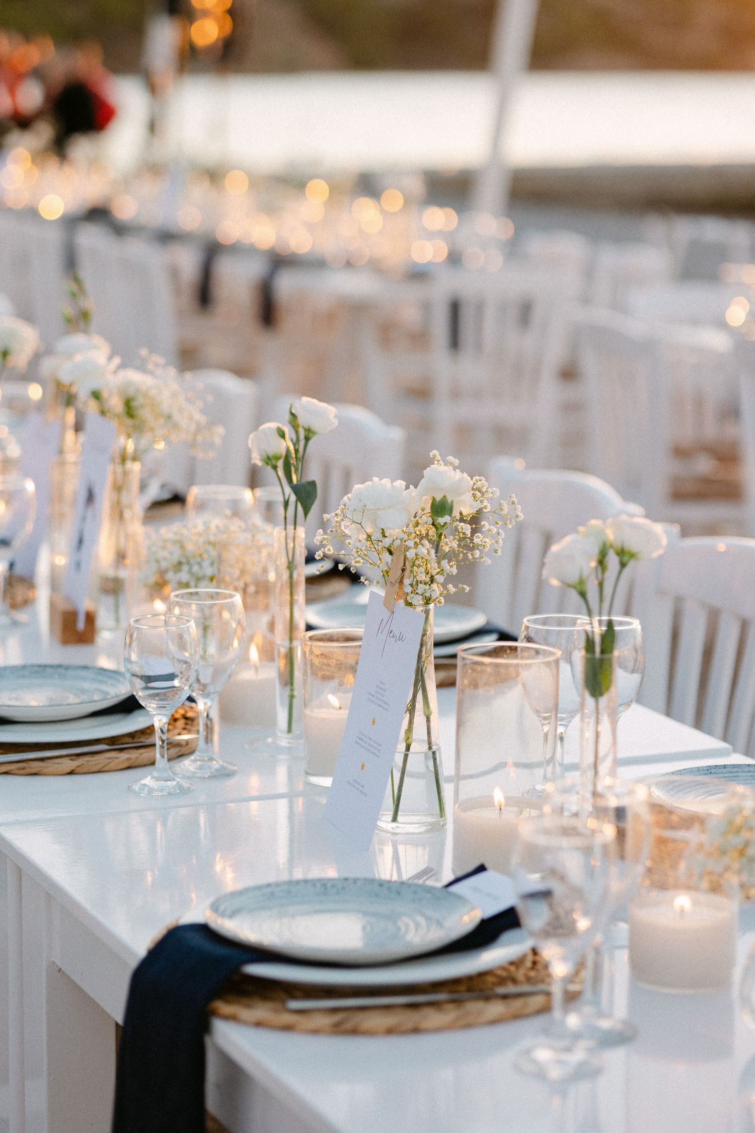 wedding table decorations showing white flowers in a vase, a lit candle, blue plates, glass wines and white chairs