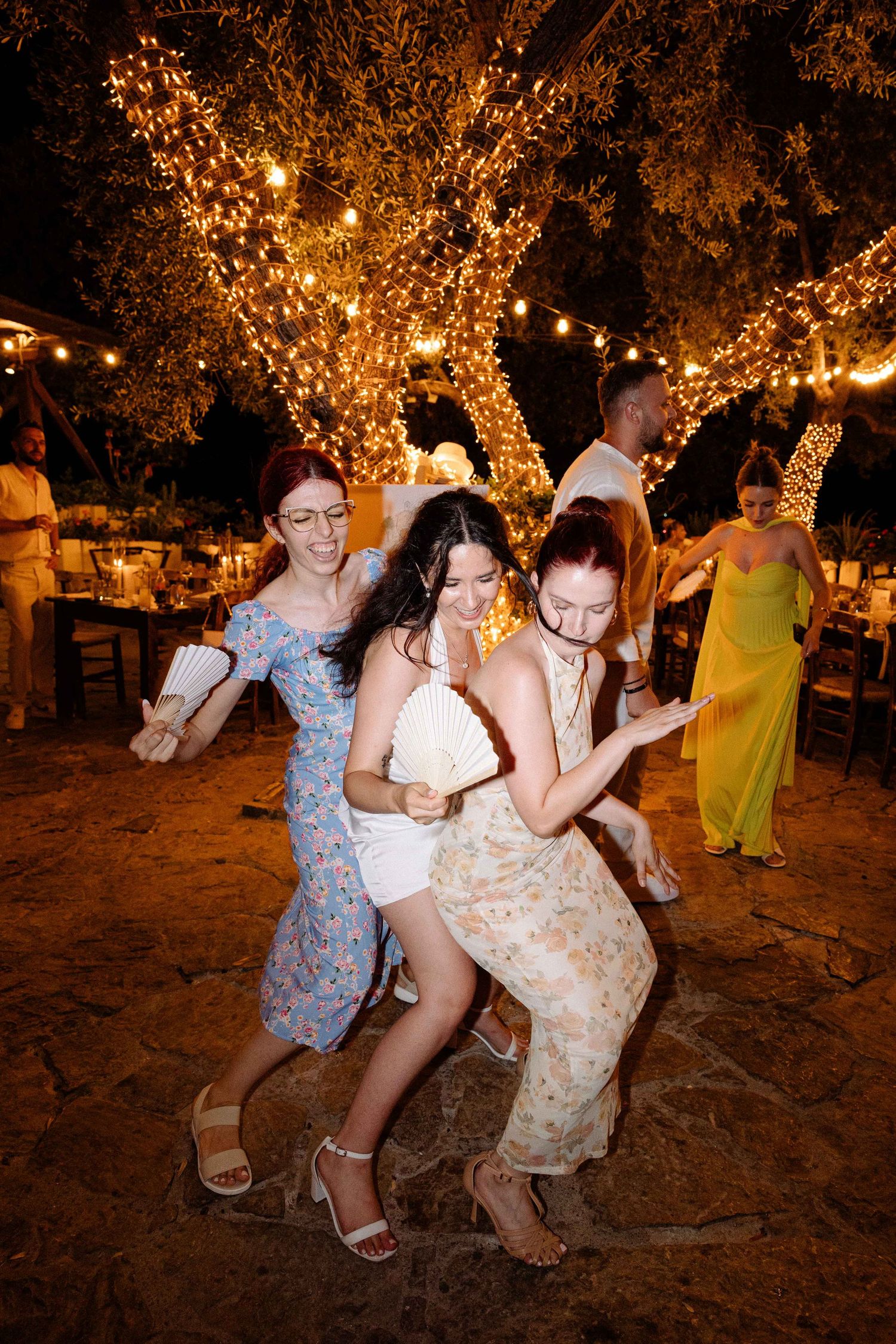 a bride and wedding guests dancing and holding fans at night surronded by trees with fairy-lights