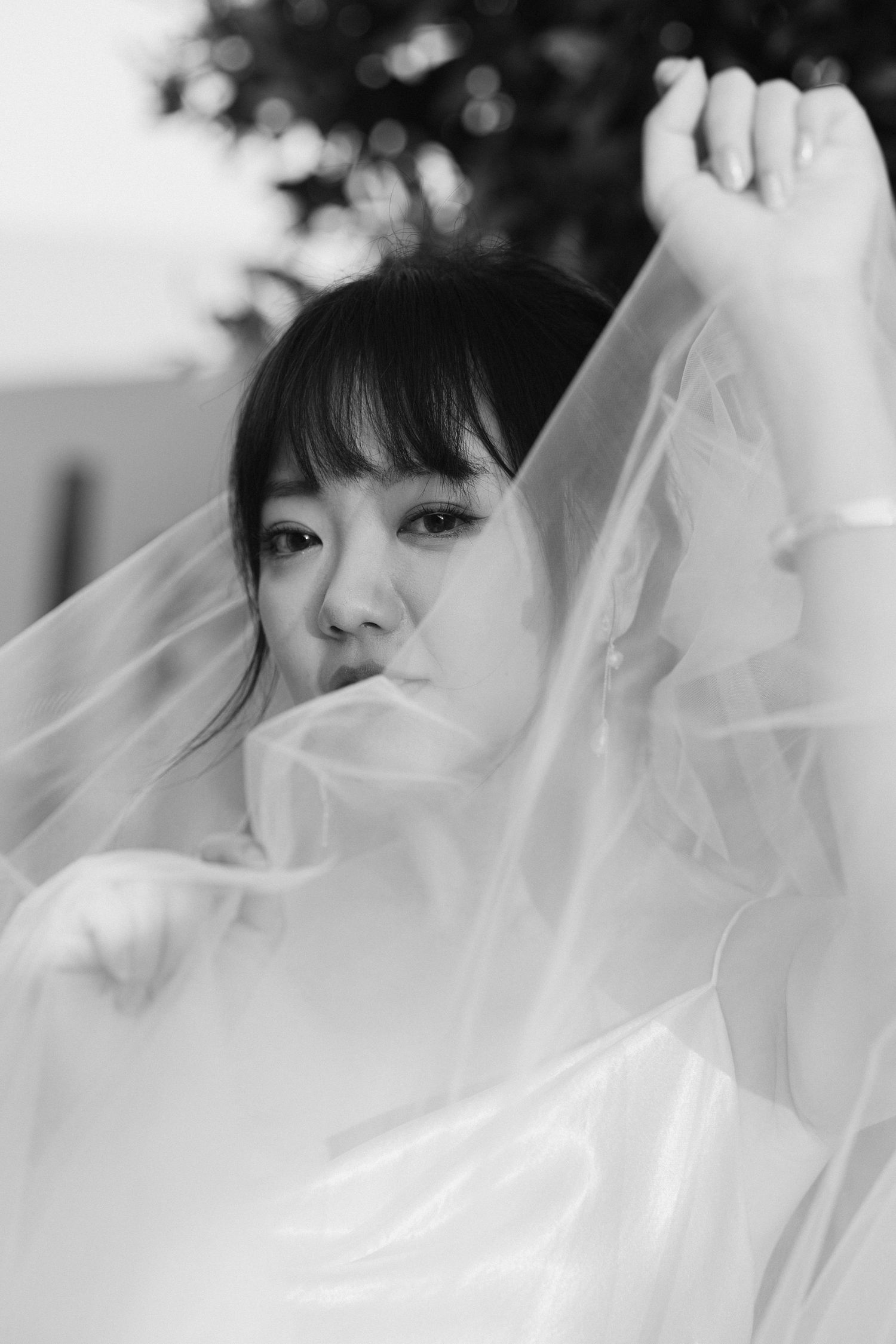 A black and white close up portrait of a bride with asian characteristics holding up her veil and looking with a soft smile at the camera