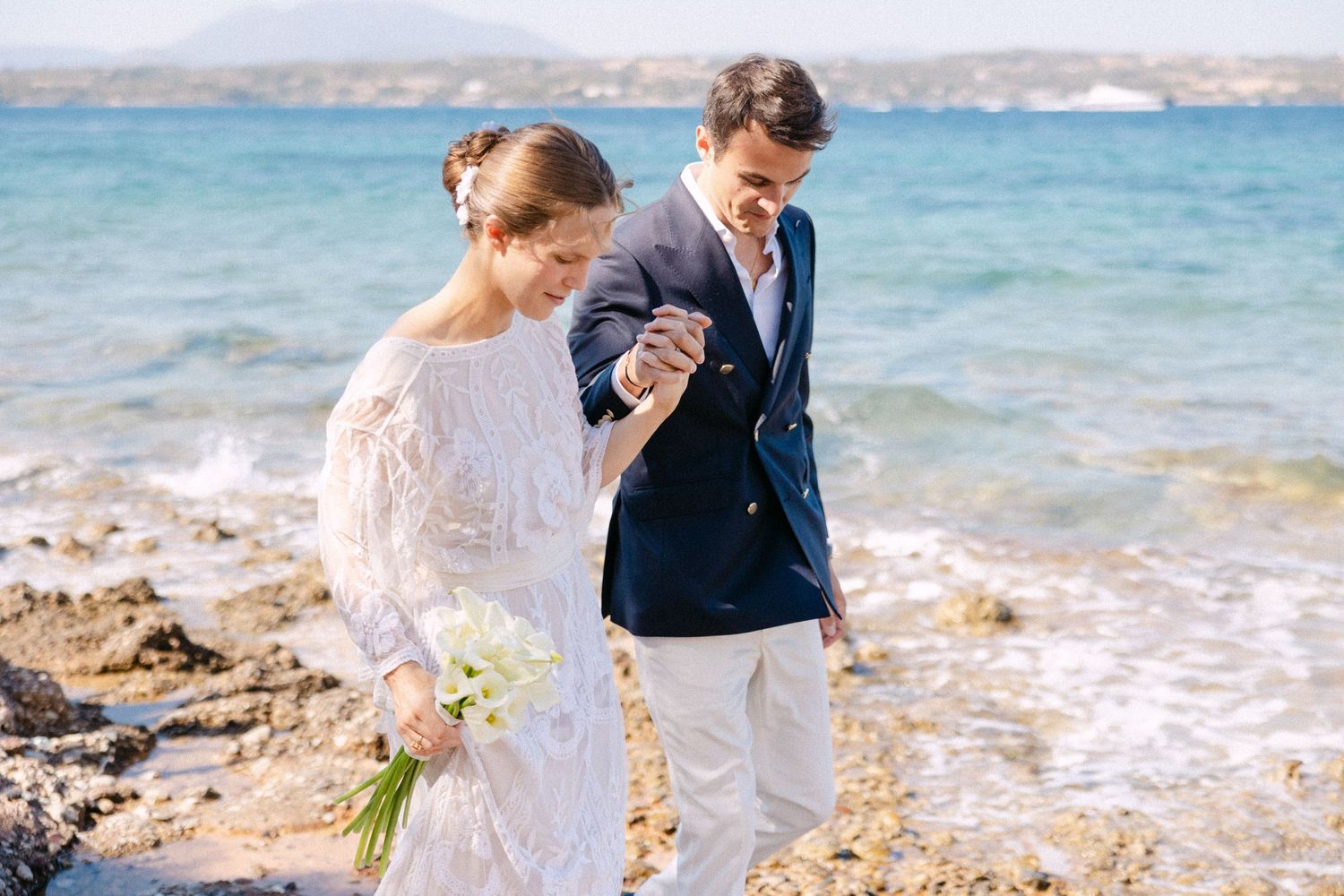 a bride and groom holding hands with the sea as a background and looking down as they are walking
