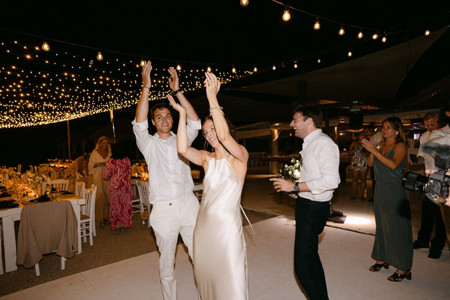 a bride and groom with their hands up in the air clapping during their wedding reception in a beach club in Spetses