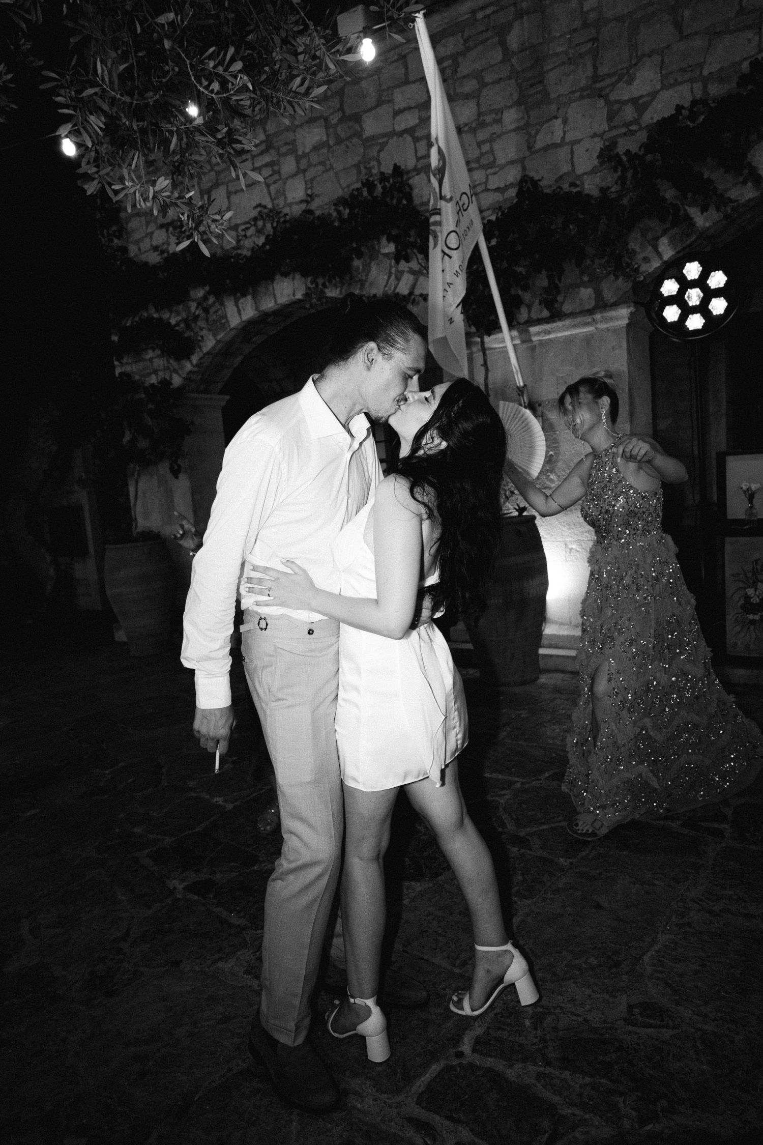 a black and white photo of a bride wearing a white mini party dress, kissing the groom on the dance floor, some people are dancing behind