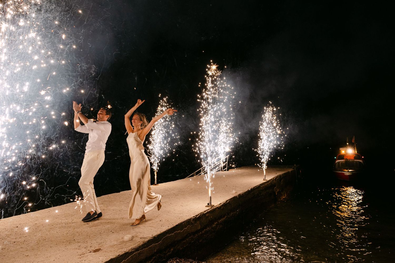 a bride and groom doing a grand entrance by a boat at sea, running in the dock with ground fireworks