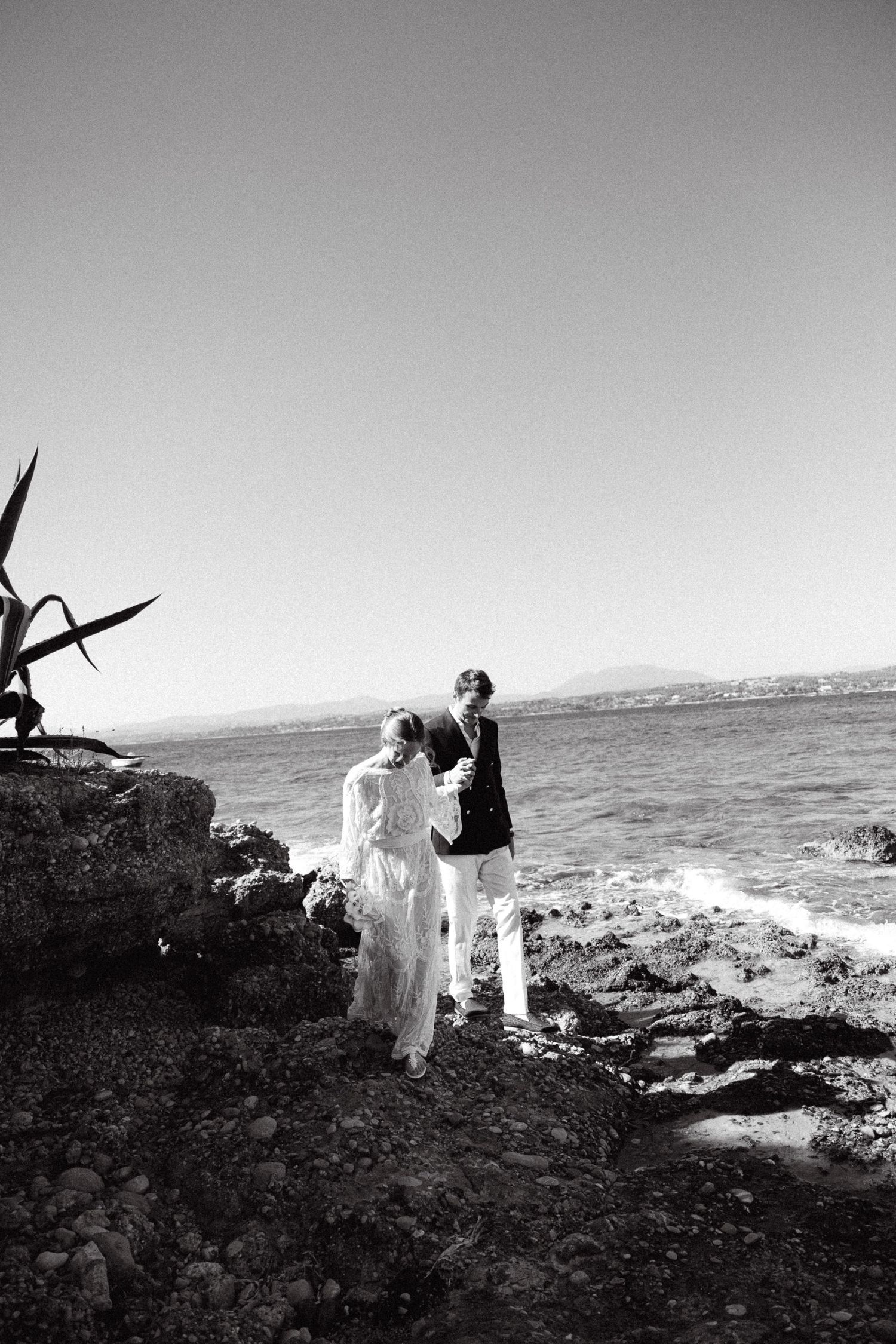 a black and white photo of a bride and groom holding hands with the sea as a background and looking down as they are walking