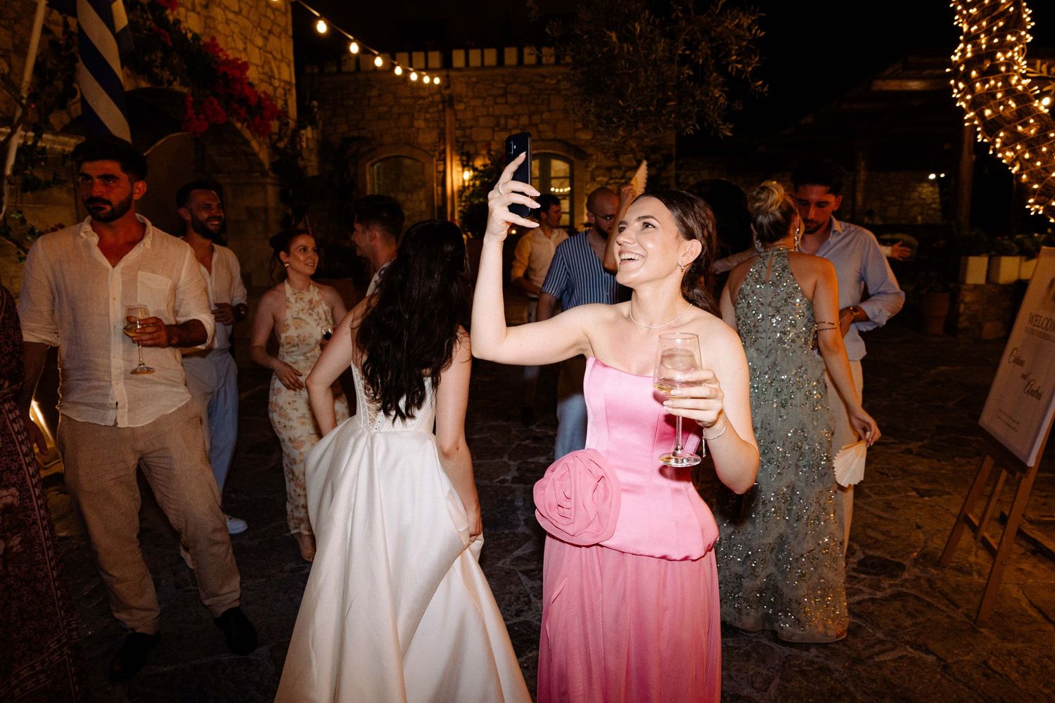 a smiling wedding guest wearing a pink dress is holding her phone taking photos amidst an outdoors party at night