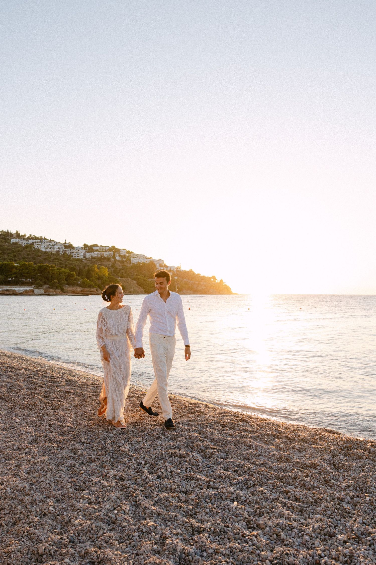 a bride and groom walking and holding hands in a beach at sunset