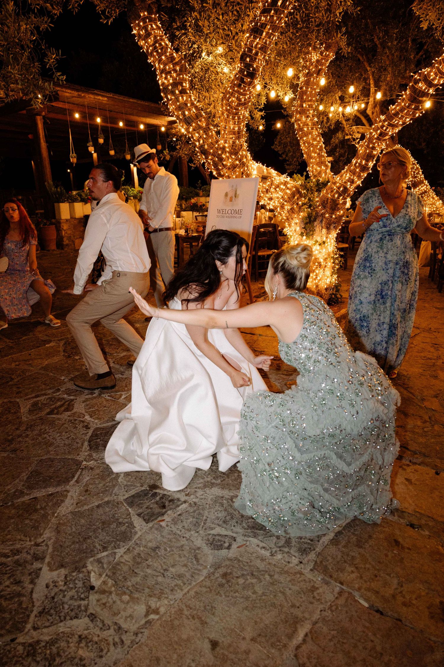 a bride dancing with her friends during an outdoors summer party, in a pose of getting low on the ground