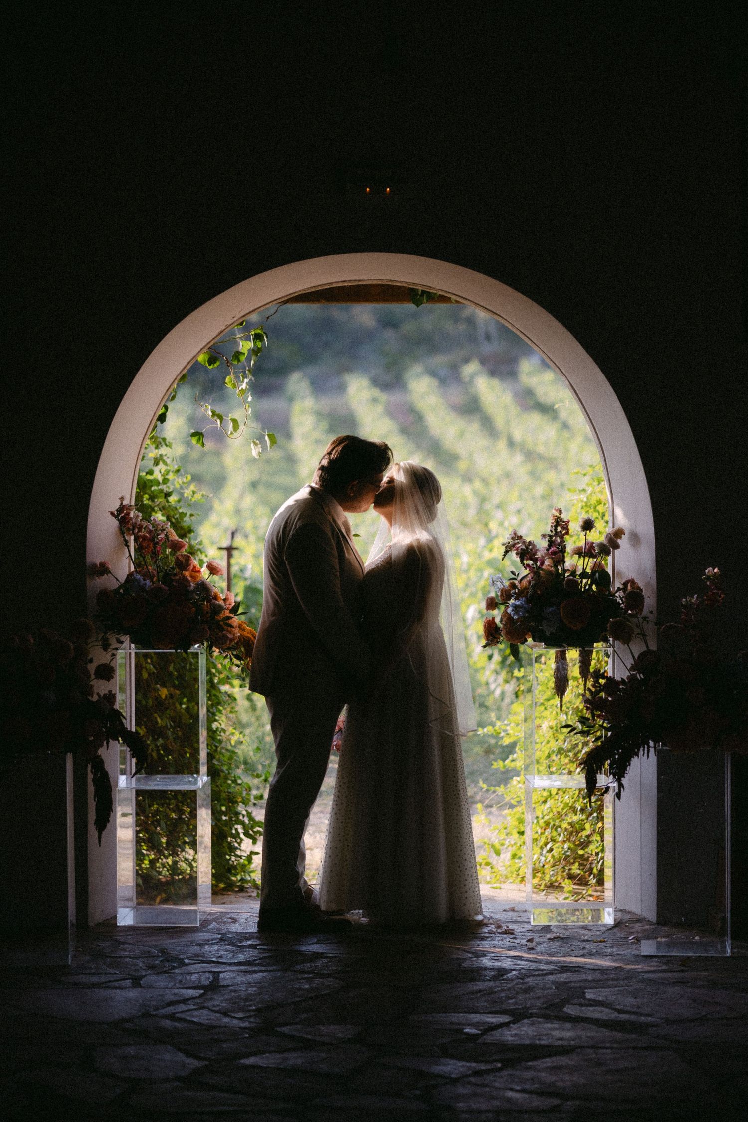 Backlit silhouette photo of a couple kissing under an arch with a vineyard as a backdrop