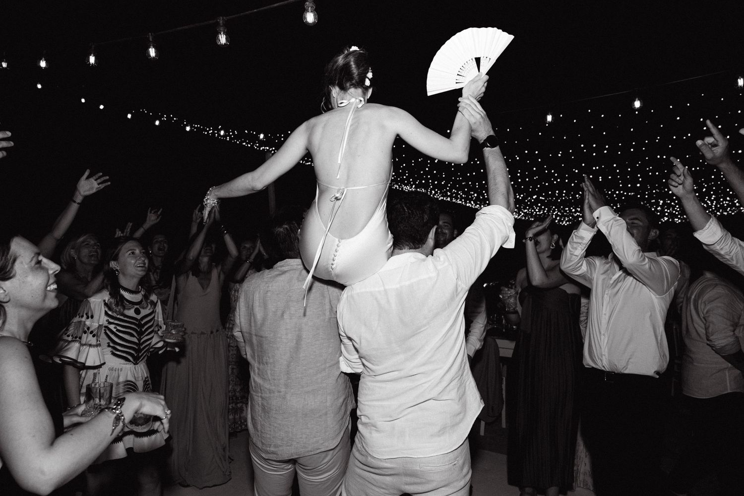 a bride sitting on two wedding guests' shoulder and dancing with her back on the camera holding a fan