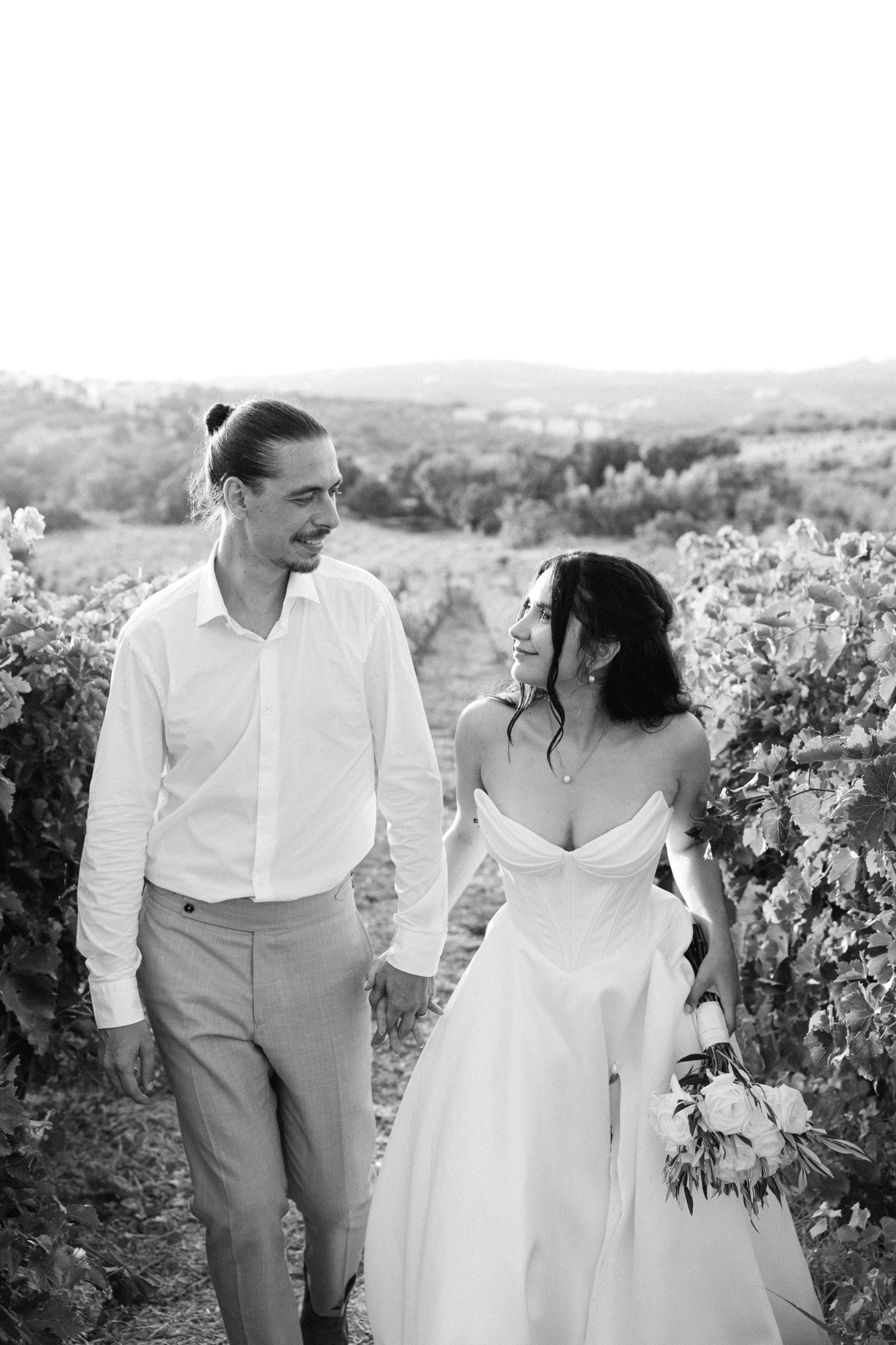 a black and white photo of a groom and a bride walking with locked arms and looking at each other among a nature field