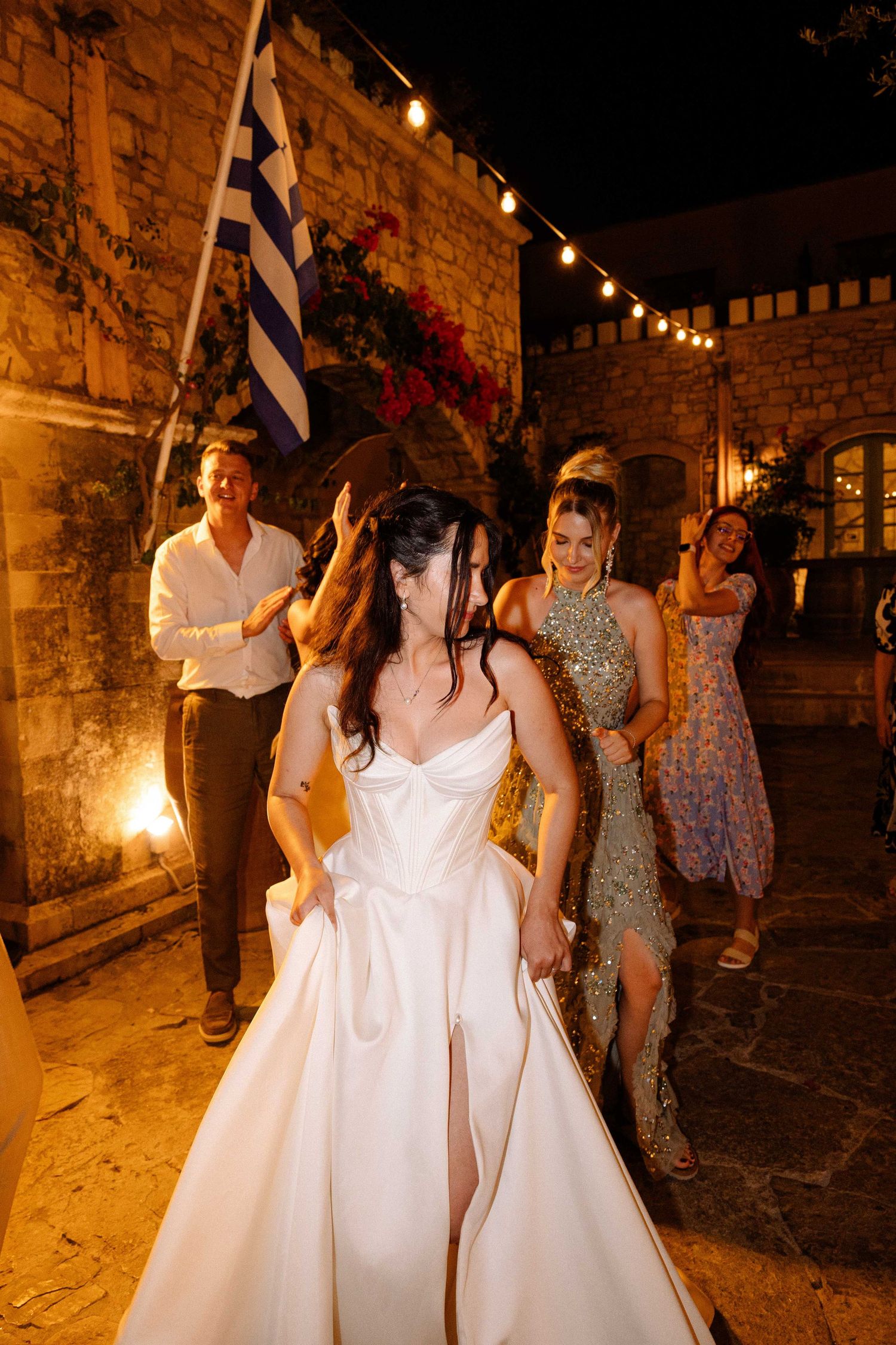 a party photo of a bride and guests dancing under outdoor warm lighting, the bride is holding her dress and a greek flag is seen behind them