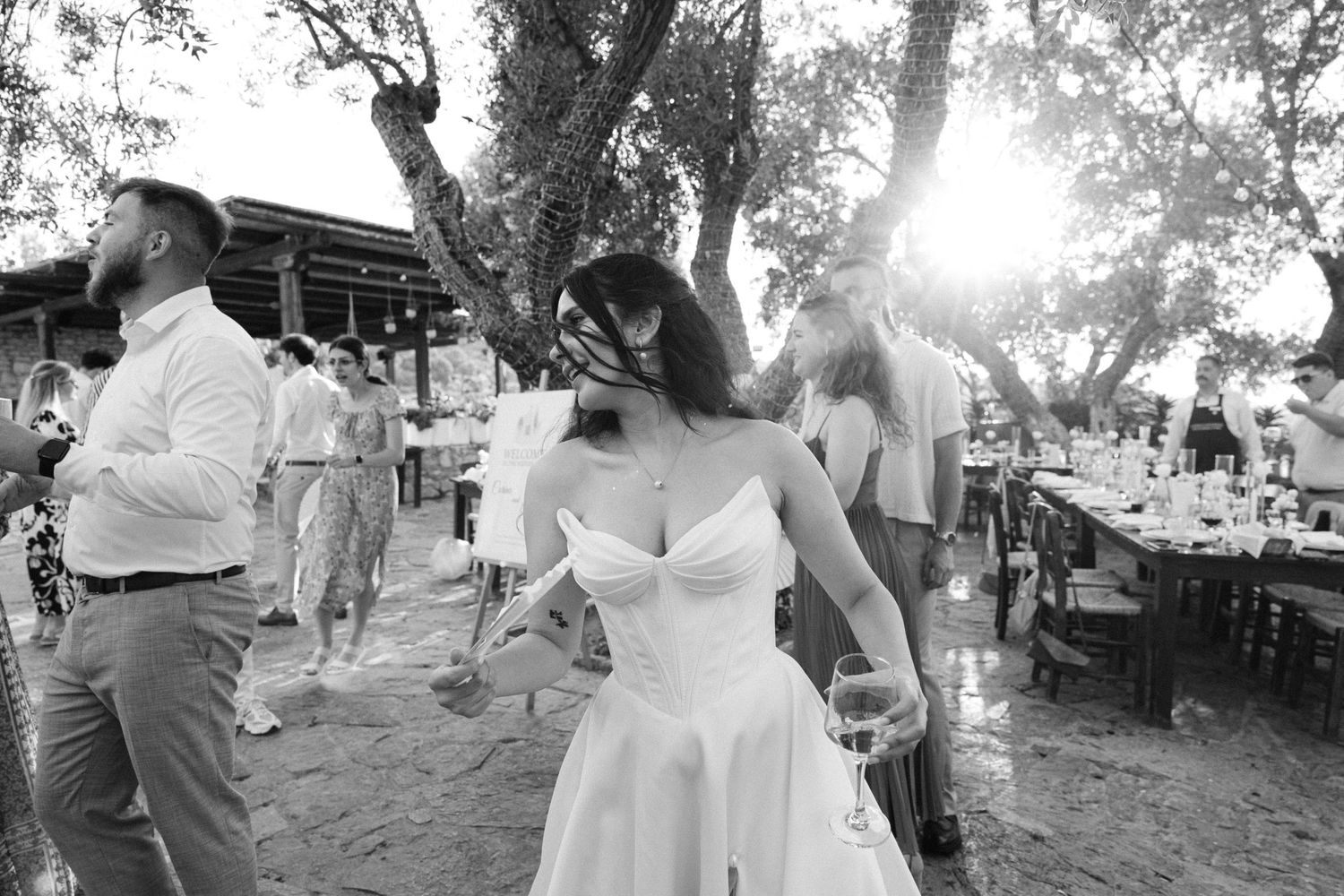 a black and white photo of a party during a wedding reception - the guests and the bride are dancing at sunset in a nature rustic setting