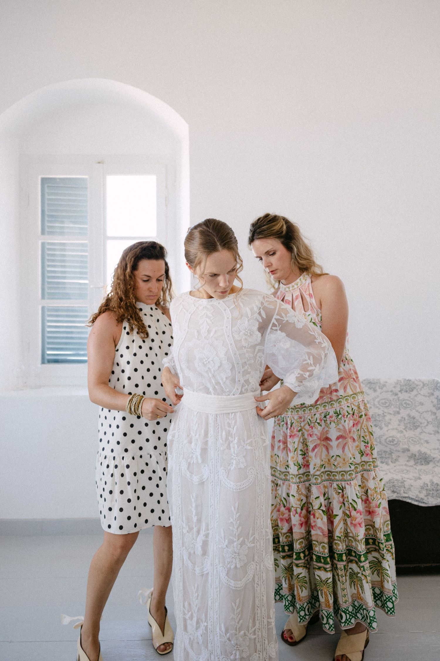 a bride in a white room being dressed by her two friends into her long-sleeved lace wedding dress - they are fixing her belt