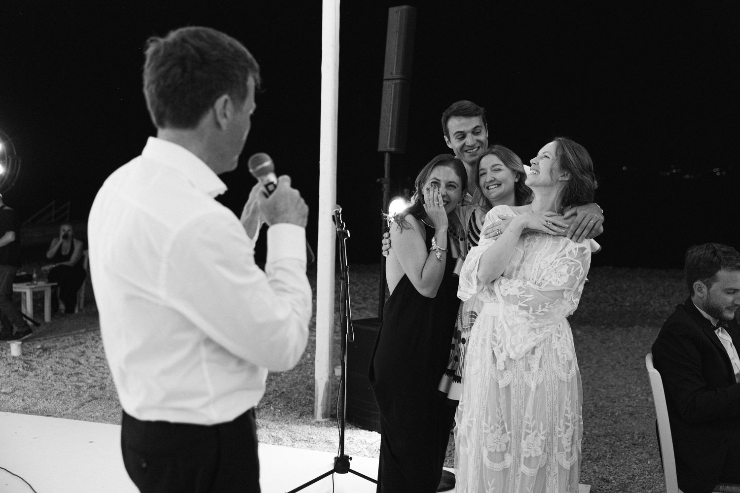 a black and white photo showing a dad reading a speech on his daughter's wedding while the bride, groom and two guests are hugging
